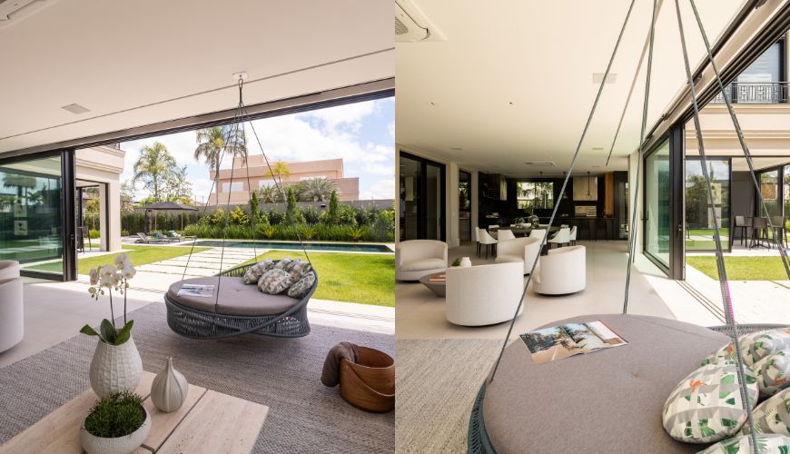 Ambiente interno e externo de uma casa luxuosa. &Agrave; esquerda, uma &aacute;rea de lazer com gramado, piscina e espregui&ccedil;adeiras, vista atrav&eacute;s de portas de vidro. No terra&ccedil;o, um balan&ccedil;o redondo cinza com almofadas estampadas e uma mesa de centro com vasos brancos. &Agrave; direita, o interior da casa com poltronas brancas e uma cozinha ao fundo