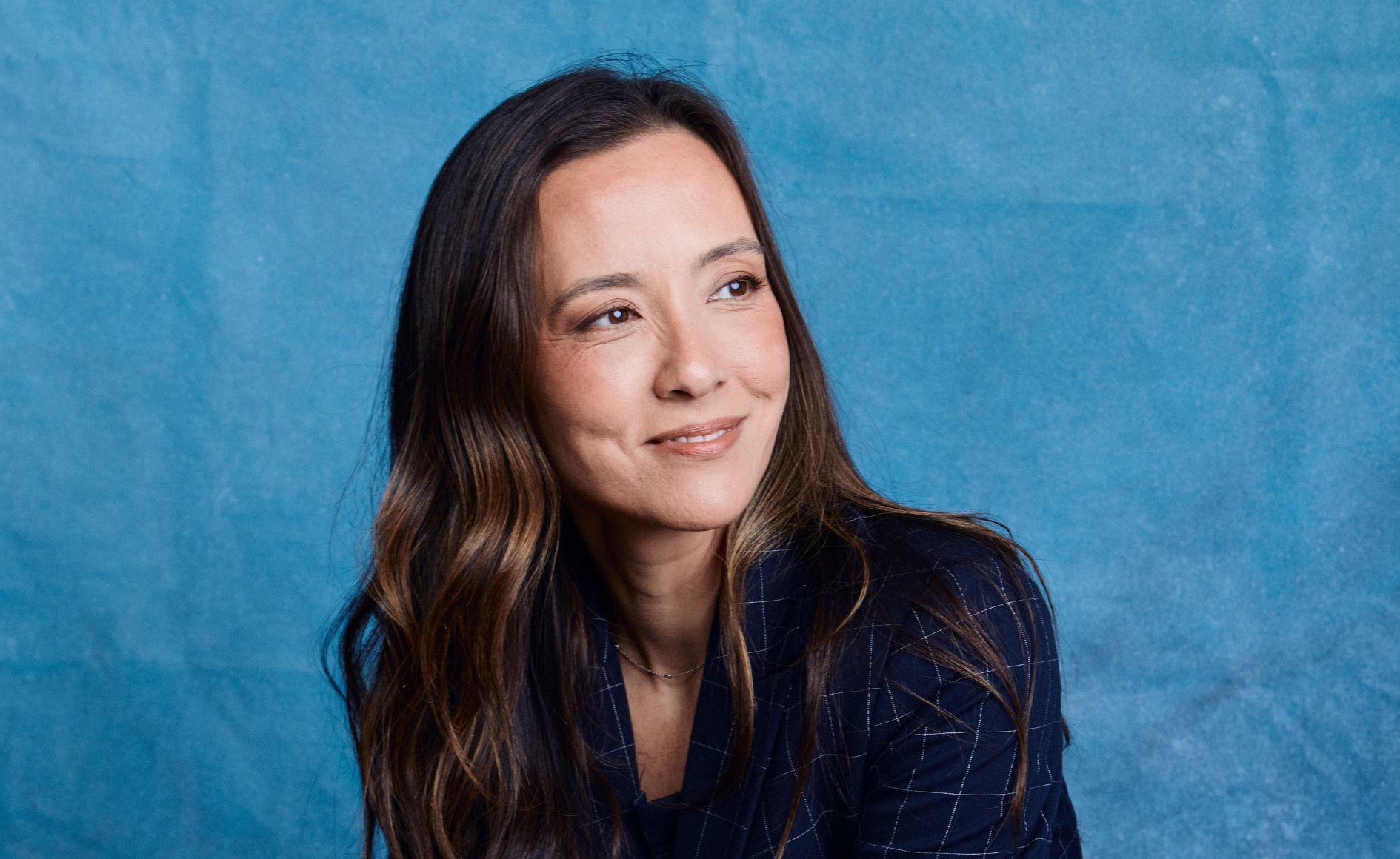 Mulher de pele clara, cabelos castanhos longos e ondulados, sorrindo e olhando para a direita, vestindo um blazer azul marinho xadrez, em fundo azul