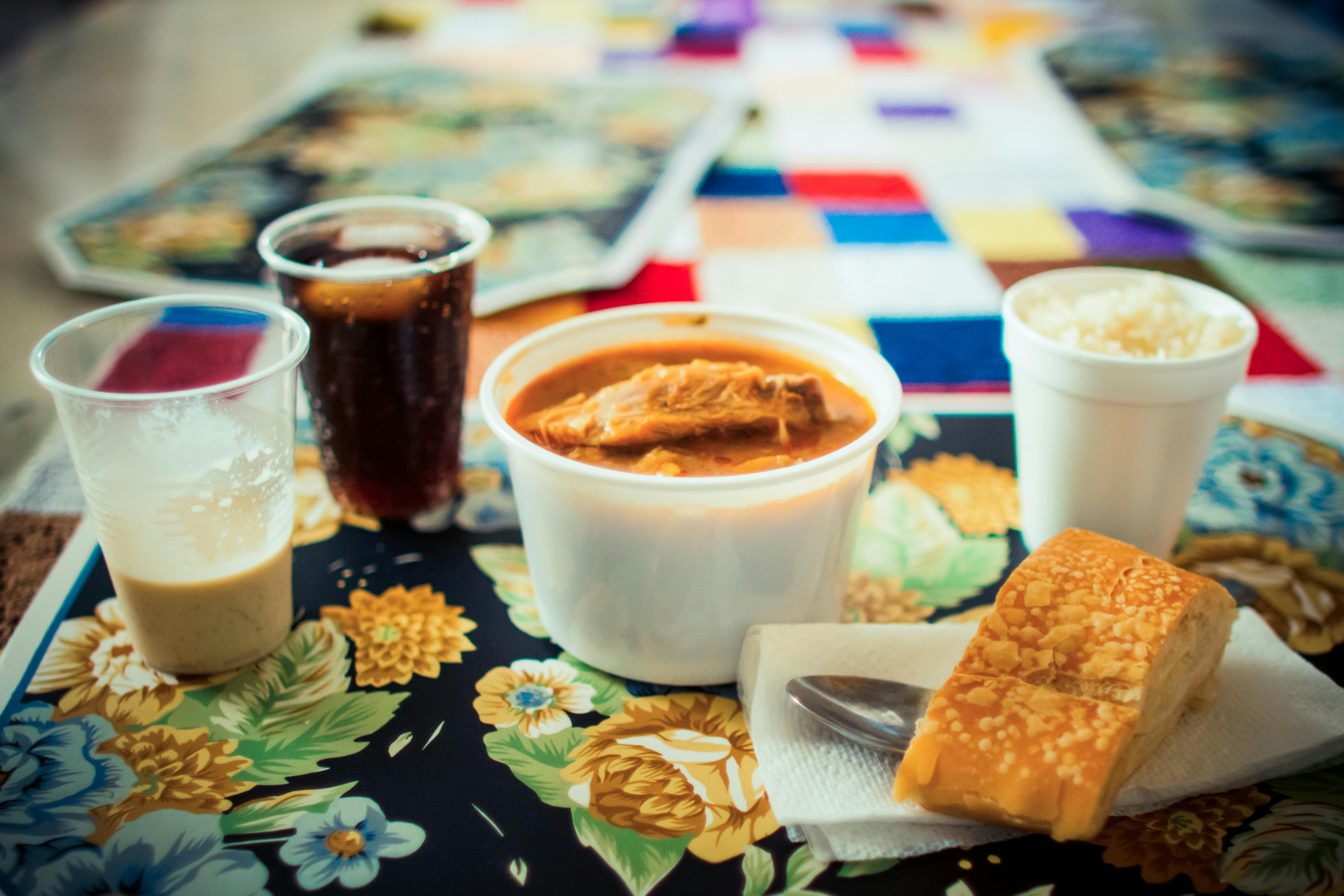 Refei&ccedil;&atilde;o simples com frango ao molho em pote branco, arroz, p&atilde;o com queijo, refrigerante e caf&eacute; em copos descart&aacute;veis, sobre toalha floral e outra de retalhos coloridos