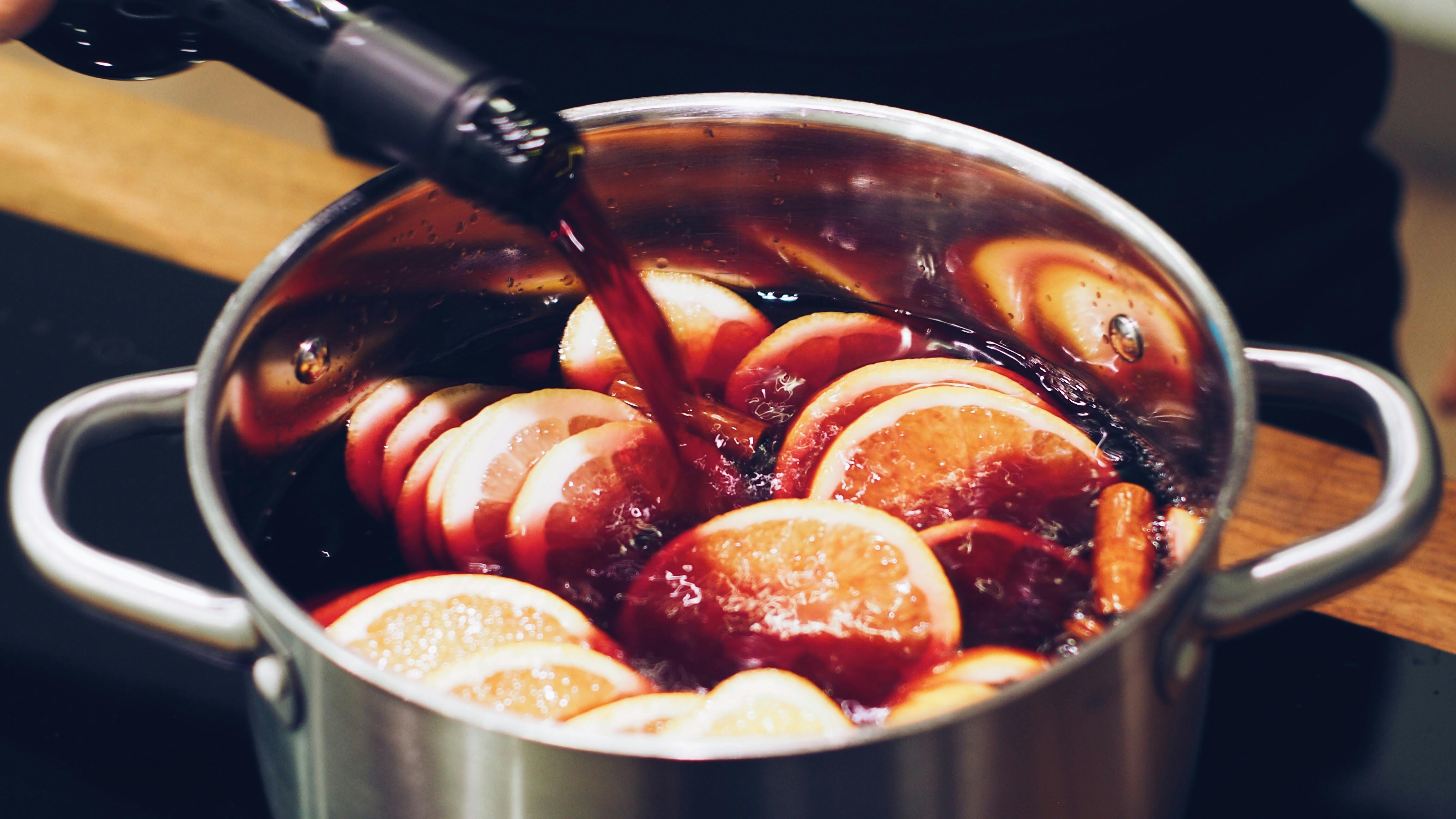 Vinho tinto sendo derramado de uma garrafa em uma panela de a&ccedil;o inoxid&aacute;vel, cheia de rodelas de laranja e especiarias, preparando uma bebida quente