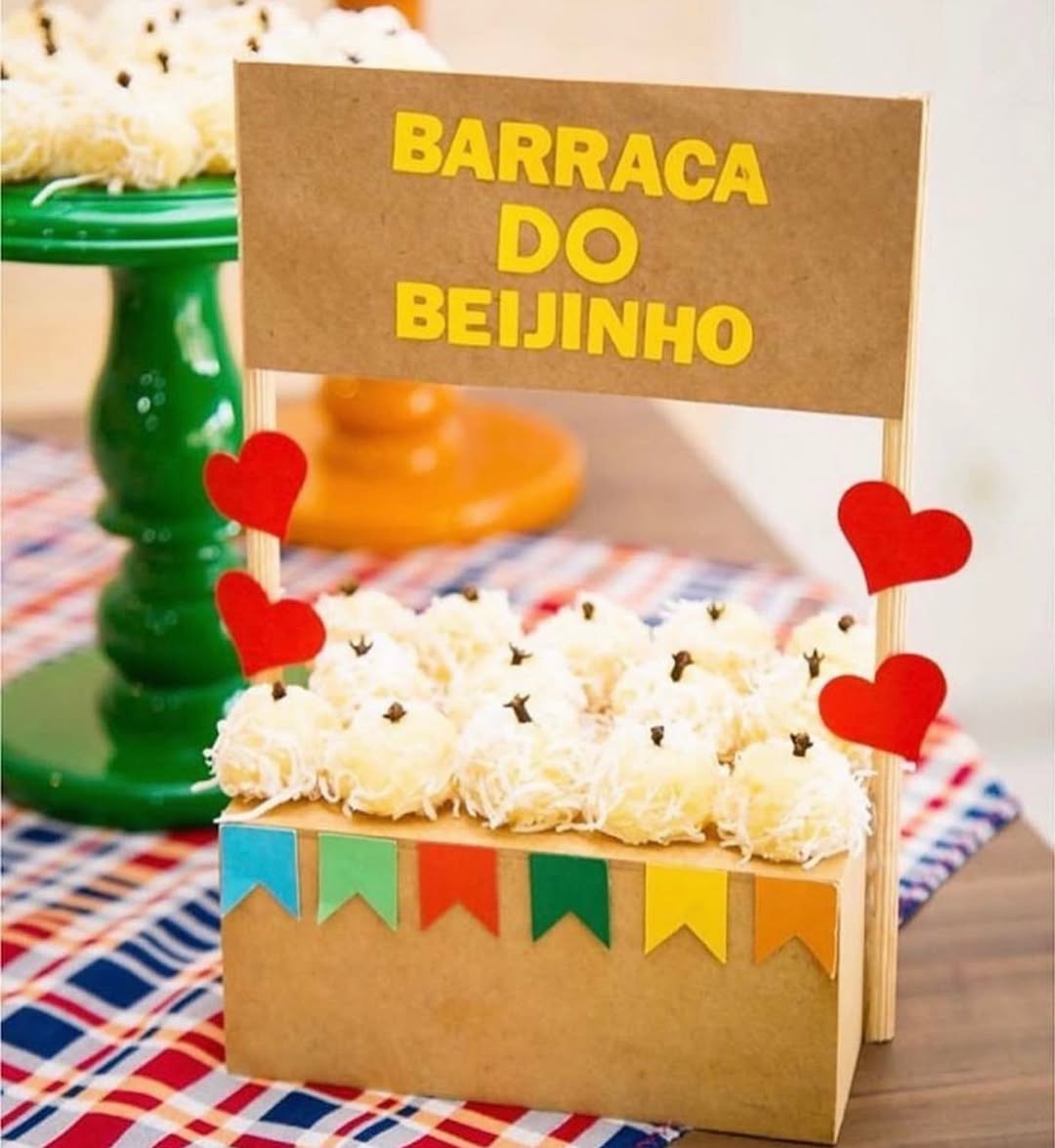 Mesa de festa junina com uma caixa de papel&atilde;o decorada com bandeirinhas coloridas, cheia de beijinhos de coco com cravo, e uma placa escrita 