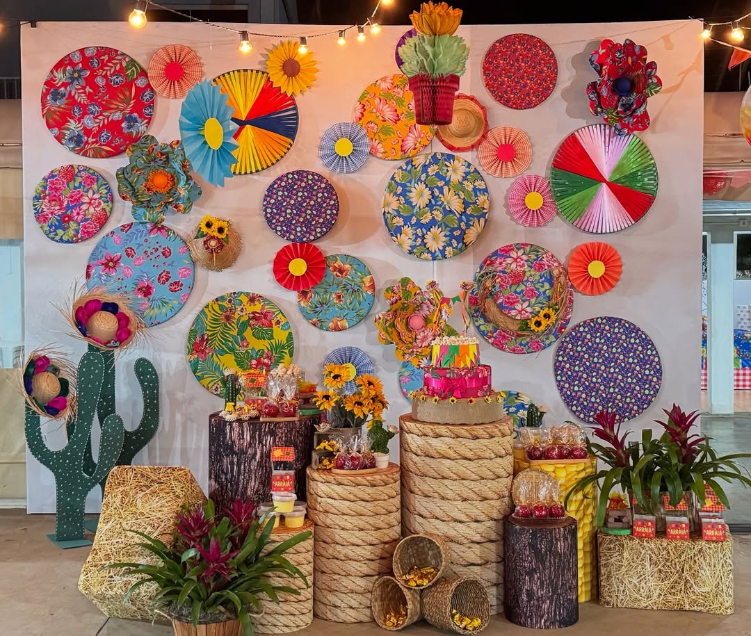 Decora&ccedil;&atilde;o de festa junina com painel branco adornado por discos de tecido floral colorido, leques de papel e chap&eacute;us de palha. Mesas r&uacute;sticas de madeira e corda exibem doces, um bolo de tr&ecirc;s andares vibrante e arranjos de girass&oacute;is, com cactos de madeira e fardos de feno complementando o cen&aacute;rio festivo