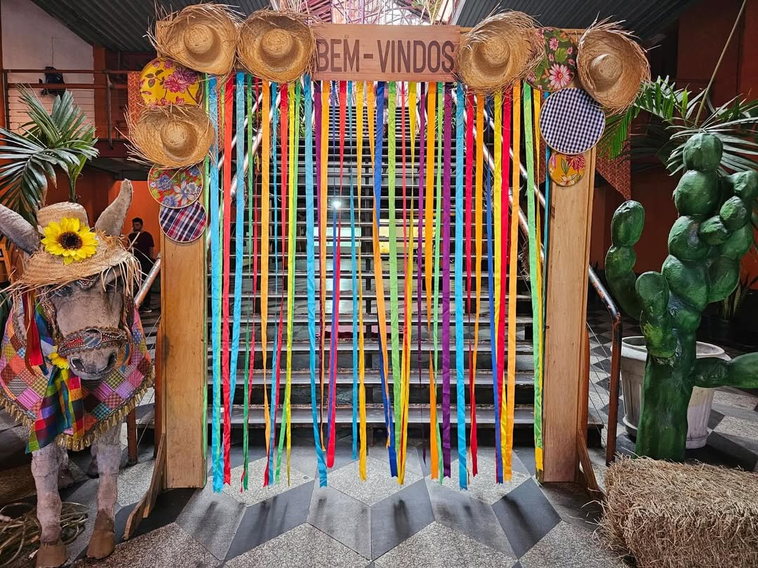 Decora&ccedil;&atilde;o de festa junina com um burro de palha usando chap&eacute;u de sol e manta colorida, ao lado de um portal de madeira com a frase 