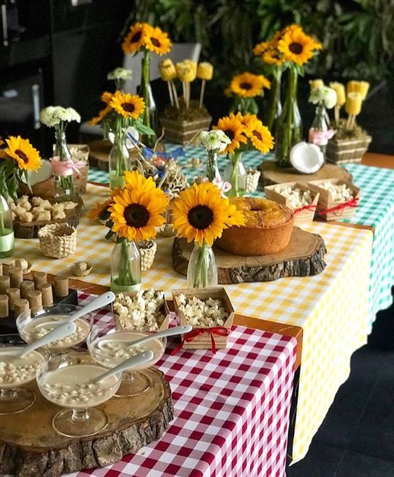Mesa de festa junina com toalhas xadrez vermelha, amarela e verde, decorada com girass&oacute;is em vasos de garrafa, bolo de fub&aacute;, pipoca em caixas de madeira, cocada, doces de milho e canjica em ta&ccedil;as de vidro
