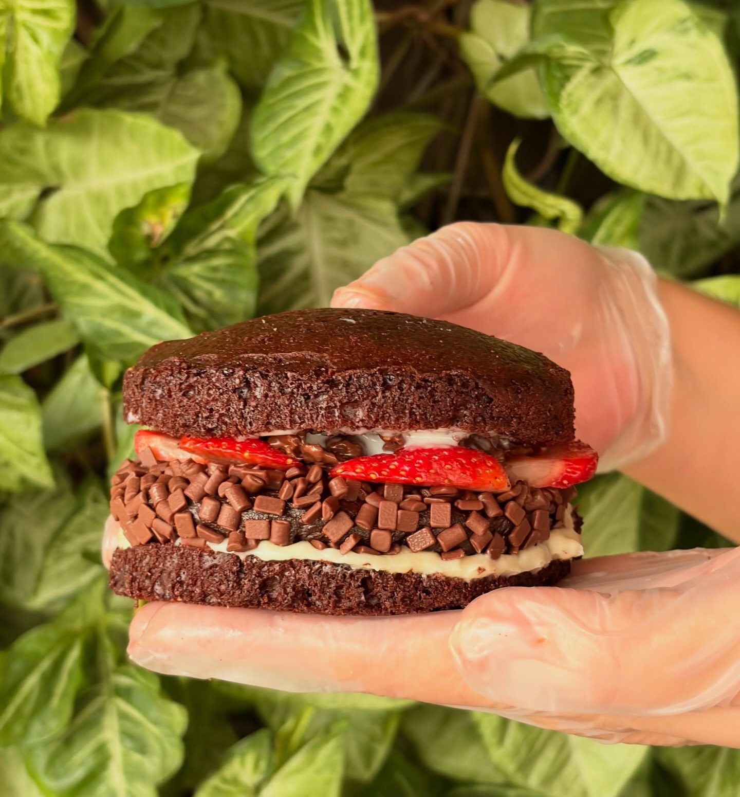 Close em um hamb&uacute;rguer doce de chocolate segurado por m&atilde;os com luvas transparentes. A sobremesa &eacute; montada com camadas de bolo de chocolate, creme branco, brigadeiro coberto por granulado e fatias de morango simulando tomates. O fundo com folhas verdes refor&ccedil;a o aspecto artesanal e chamativo da cria&ccedil;&atilde;o viral.