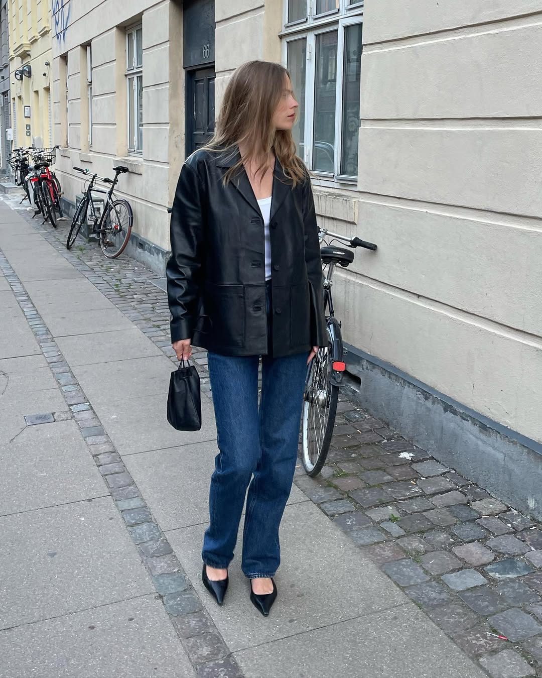 Foto de uma mulher loira usando um look composto por jaqueta de couro preta, regata branca, cal&ccedil;a jeans azul e salto preto