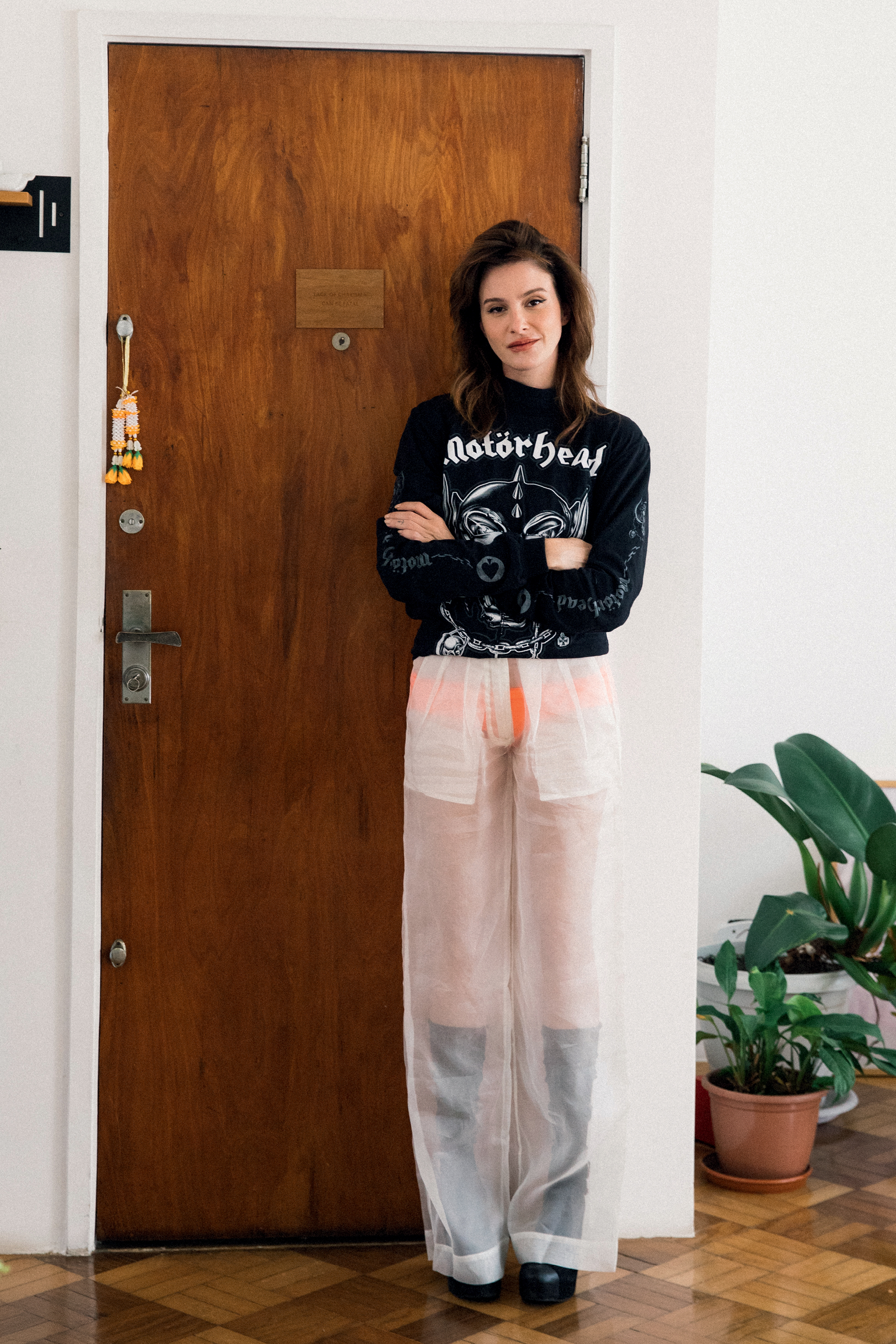 Foto de Verena Figueiredo. Nela, ela aparece em frente a uma porta de madeira. Ela usa blusa preta, cal&ccedil;a transparente e bota de couro