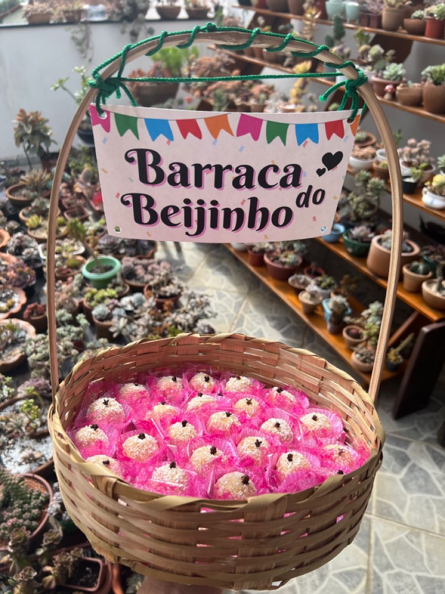 Cesta de bambu com beijinhos de coco em forminhas cor-de-rosa, cada um com um cravo no topo. Um cartaz de festa junina escrito 
