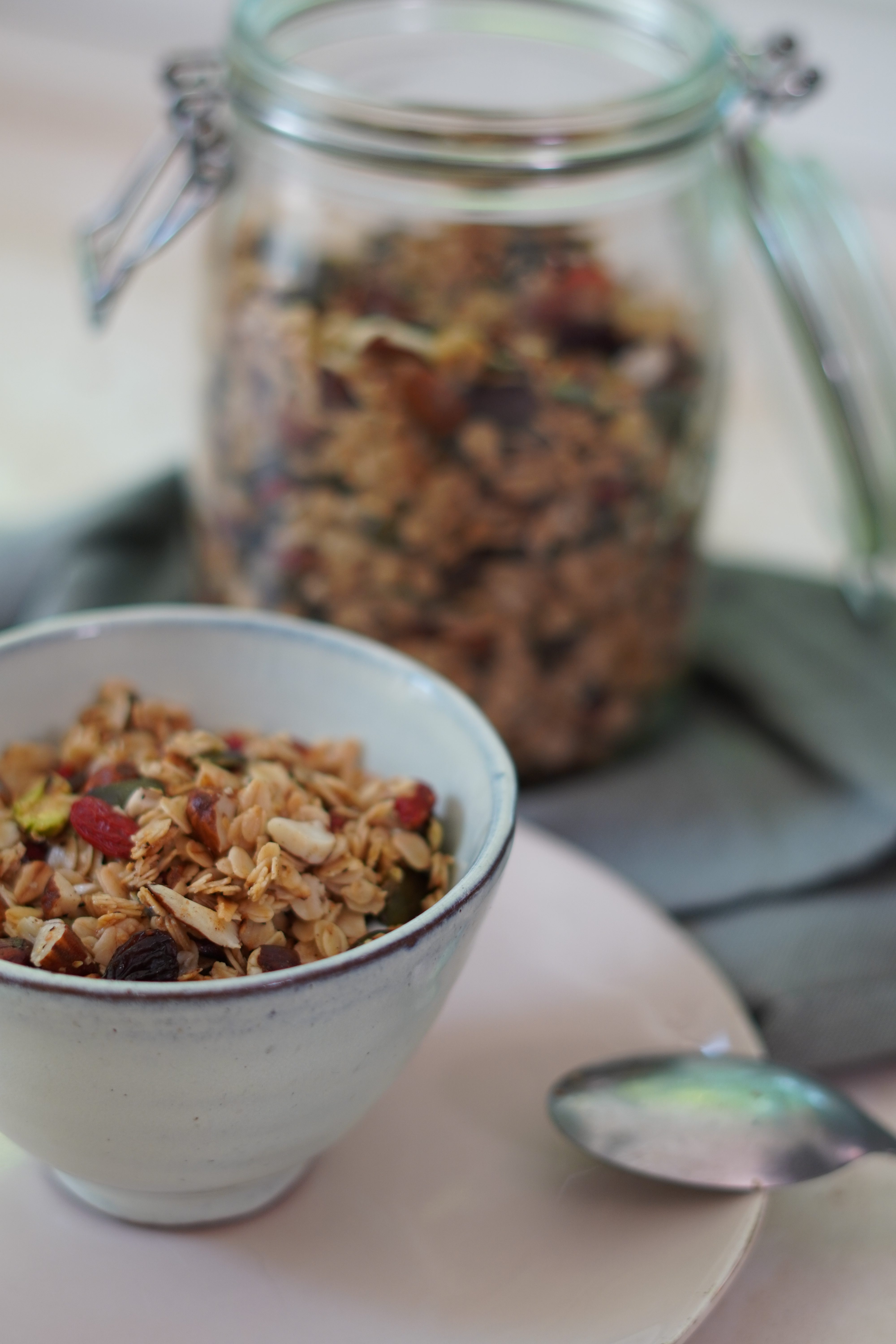Uma tigela de granola caseira com aveia, nozes, sementes e frutas secas, como goji berries, em primeiro plano. Ao fundo, um pote de vidro grande cheio de granola, sobre um pano cinza. Uma colher de metal est&aacute; em um prato rosa claro ao lado da tigela