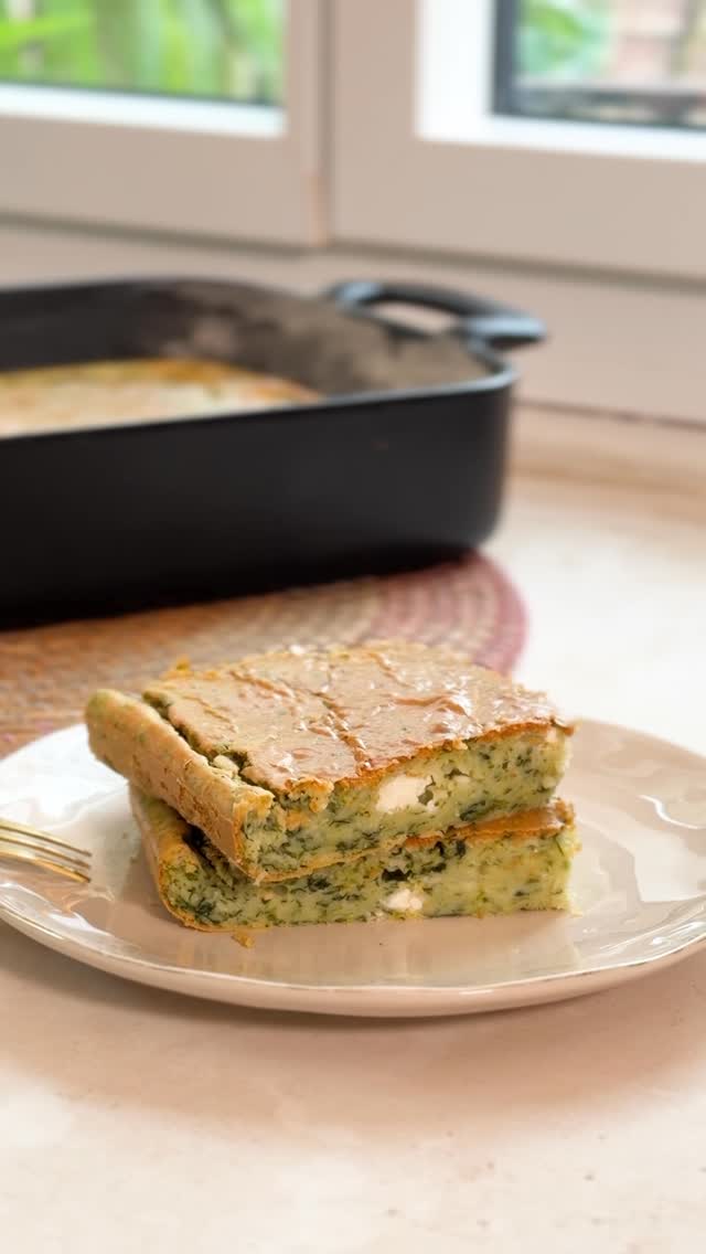 Duas fatias de torta salgada verde com peda&ccedil;os de queijo branco, empilhadas em um prato claro, com uma assadeira preta ao fundo