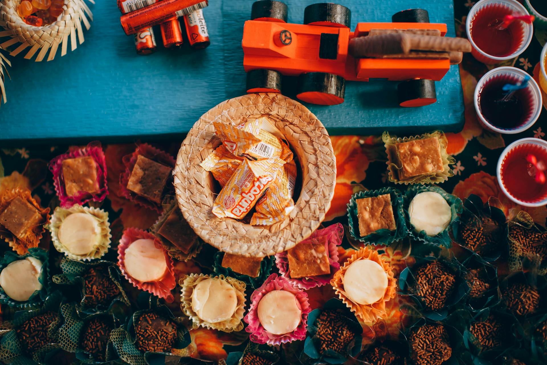 Mesa de festa junina com doces variados, como pa&ccedil;ocas em um chap&eacute;u de palha, brigadeiros, beijinhos e doces de amendoim, al&eacute;m de copos com bebidas vermelhas e um brinquedo de madeira laranja, sobre uma toalha florida