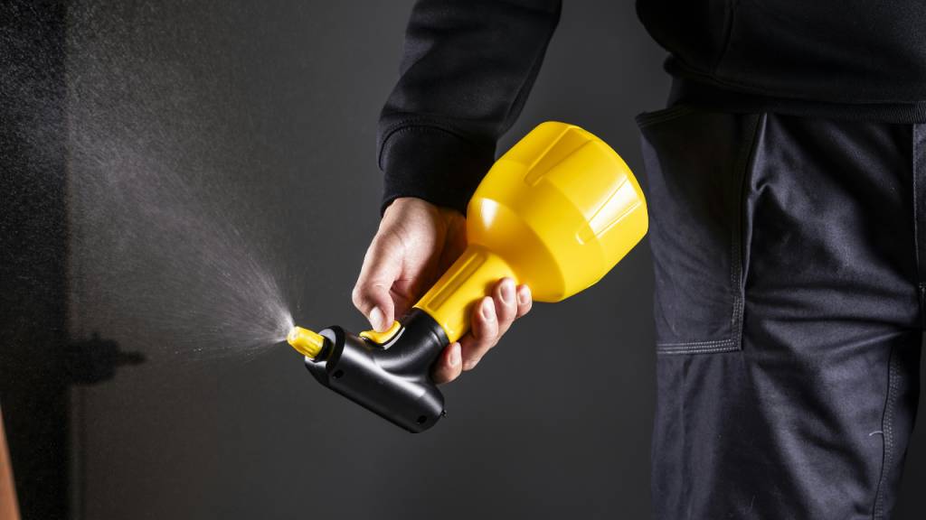 Uma imagem em plano detalhe e em ambiente interno mostra as m&atilde;os e o tronco de uma pessoa utilizando um pulverizador manual. A pessoa veste roupas escuras &mdash; um casaco de manga comprida preto e cal&ccedil;as de tecido grosso cinza-escuro. Ela est&aacute; posicionada do centro para o lado direito da imagem, de perfil parcial. Com a m&atilde;o direita, a pessoa segura o pulverizador. O equipamento &eacute; composto por um reservat&oacute;rio pl&aacute;stico **amarelo vibrante** na parte superior (com formato c&ocirc;nico invertido), acoplado a um cabo preto ergon&ocirc;mico na parte inferior, semelhante ao cabo de uma ferramenta ou furadeira. O polegar da pessoa est&aacute; posicionado sobre um gatilho amarelo no cabo. A ponta do pulverizador possui um bico dosador amarelo direcionado para o lado esquerdo da imagem. Dele, sai uma **n&eacute;voa fina e esbranqui&ccedil;ada de spray**, que se espalha pelo ar em dire&ccedil;&atilde;o a uma superf&iacute;cie escura e indefinida no canto esquerdo. O fundo da imagem &eacute; completamente escuro e plano, o que faz com que a cor amarela do pulverizador e a n&eacute;voa do spray se destaquem com nitidez.