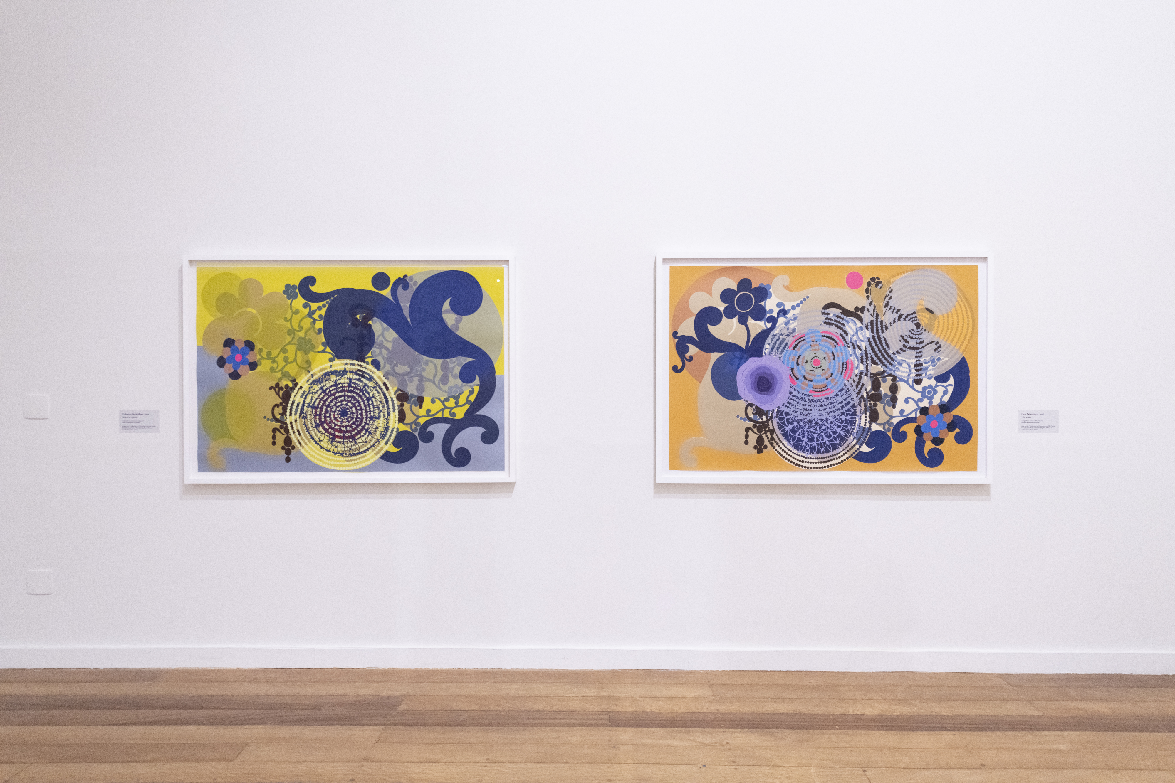 Duas obras de arte abstratas emolduradas, com fundos amarelo e laranja, exibem padr&otilde;es circulares e florais em tons de azul, roxo e cinza, penduradas em uma parede branca de galeria com piso de madeira