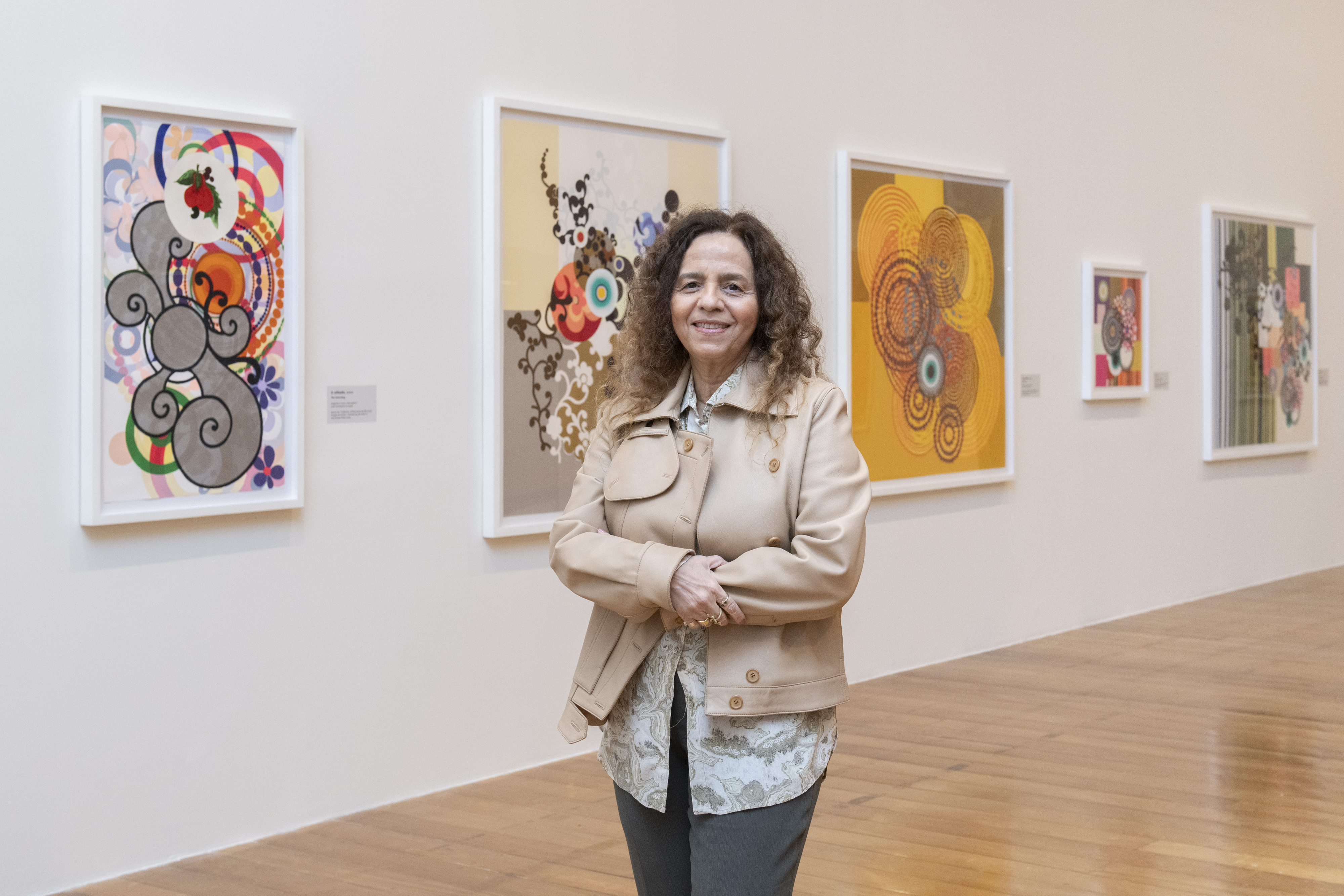 Mulher de cabelos cacheados castanhos, sorrindo, vestindo jaqueta bege e camisa estampada, bra&ccedil;os cruzados, em uma galeria de arte com quadros abstratos coloridos na parede branca