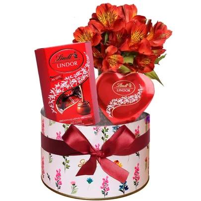Uma caixa redonda branca com estampa floral colorida, la&ccedil;o de fita vermelho, contendo um buqu&ecirc; de flores alstroem&eacute;rias laranjas, uma caixa vermelha de chocolates Lindt Lindor ao leite e uma caixa de bombons Lindt Lindor em formato de cora&ccedil;&atilde;o
