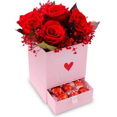Caixa rosa com buqu&ecirc; de rosas vermelhas e flores secas. Na frente, um cora&ccedil;&atilde;o vermelho e as palavras Amo Voc&ecirc;. Uma gaveta inferior est&aacute; aberta, revelando bombons de chocolate embrulhados em papel vermelho e dourado