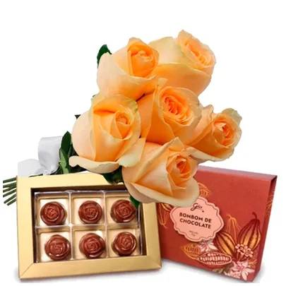 Buqu&ecirc; de seis rosas cor p&ecirc;ssego com folhas verdes e la&ccedil;o branco, ao lado de uma caixa dourada aberta com seis bombons em formato de rosa e uma caixa de bombons vermelha com ilustra&ccedil;&atilde;o de cacau e o texto Bombom de Chocolate