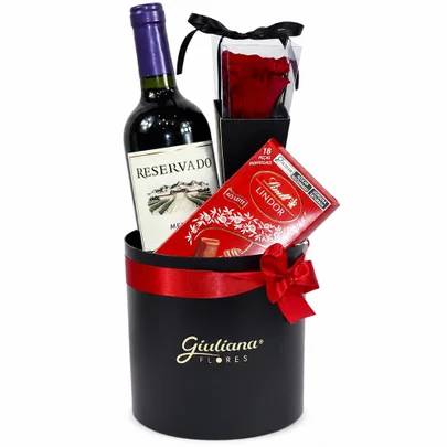 Cesta de presente preta com fita vermelha e la&ccedil;o, contendo uma garrafa de vinho tinto Reservado, uma caixa de bombons Lindt Lindor ao leite e uma rosa vermelha em embalagem transparente