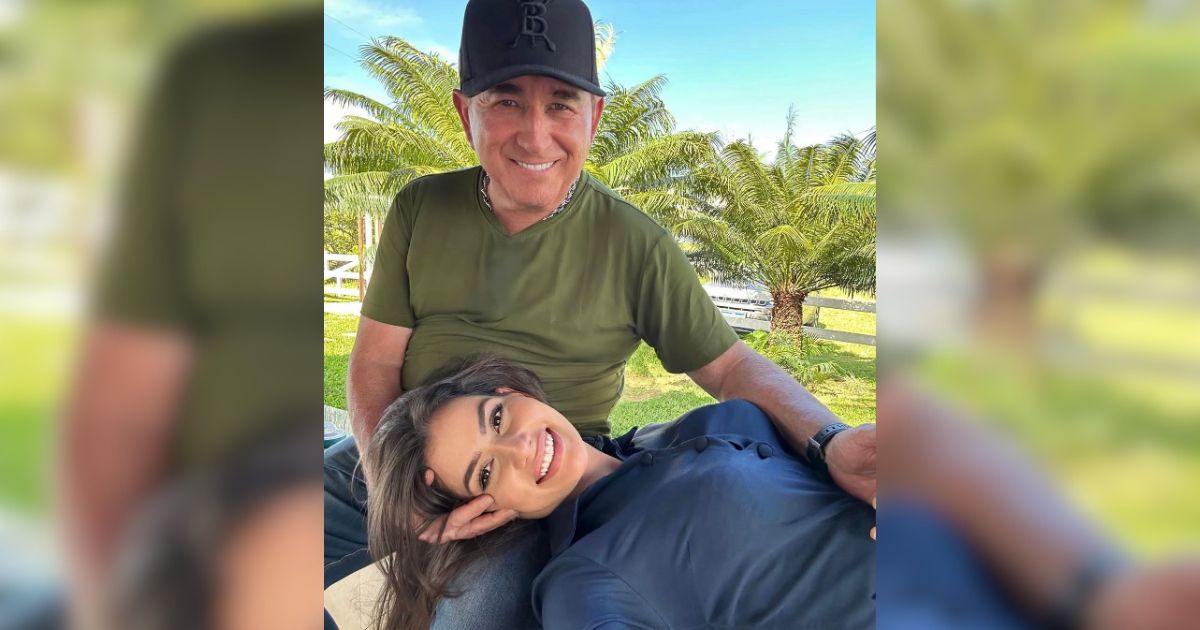 Um homem sorridente de bon&eacute; preto e camiseta verde-oliva sentado, com uma mulher de blusa azul-marinho deitada em seu colo, ambos sorrindo para a c&acirc;mera em um ambiente externo com palmeiras e c&eacute;u azul