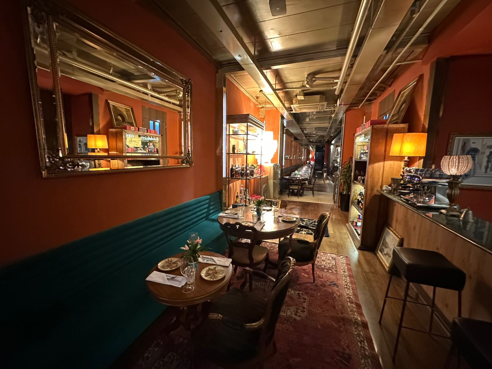Interior de restaurante com paredes cor de telha, espelho grande, e um longo banco estofado verde-azulado. Mesas redondas de madeira com cadeiras escuras, tapete persa no ch&atilde;o, e um balc&atilde;o com lumin&aacute;rias e garrafas. Ao fundo, mais mesas iluminadas, criando um ambiente aconchegante e intimista