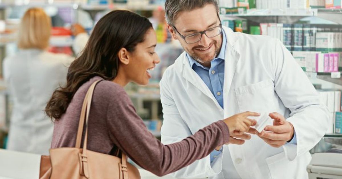 Farmac&ecirc;utico sorridente de &oacute;culos e jaleco branco entrega um pequeno pacote a uma cliente jovem, que aponta para o produto com express&atilde;o feliz, em uma farm&aacute;cia com prateleiras cheias de medicamentos ao fundo