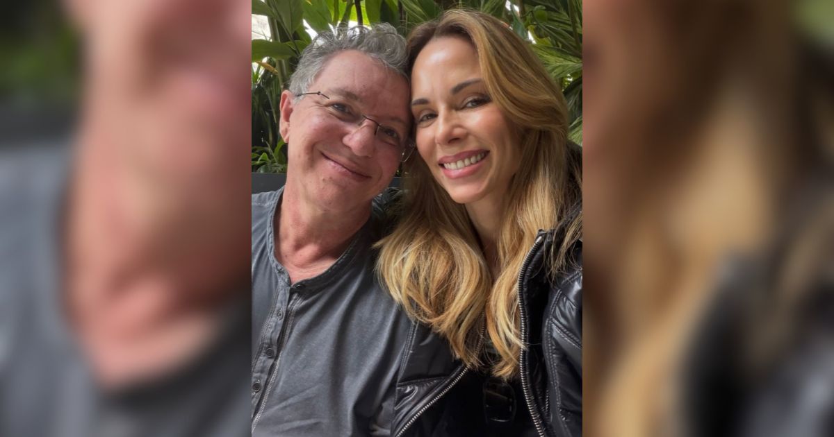 Um homem de &oacute;culos e cabelo grisalho sorri ao lado de uma mulher loira com jaqueta preta, ambos com express&otilde;es felizes, em um ambiente externo com folhagem verde ao fundo