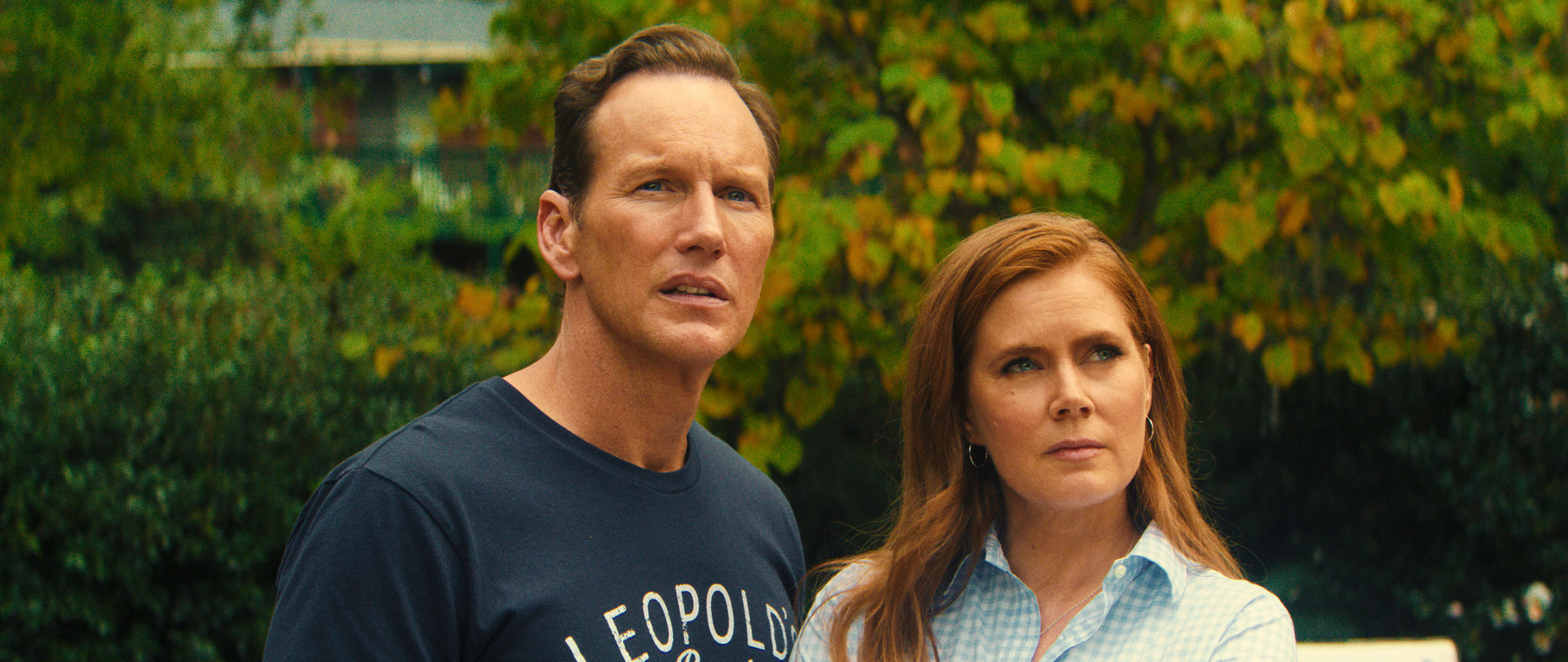 Patrick Wilson e Amy Adams, com express&otilde;es s&eacute;rias e preocupadas, olham para o alto e para a direita. Ele veste uma camiseta azul-marinho, e ela, uma camisa xadrez azul clara, com cabelos ruivos soltos. Ao fundo, folhagem verde e amarela.