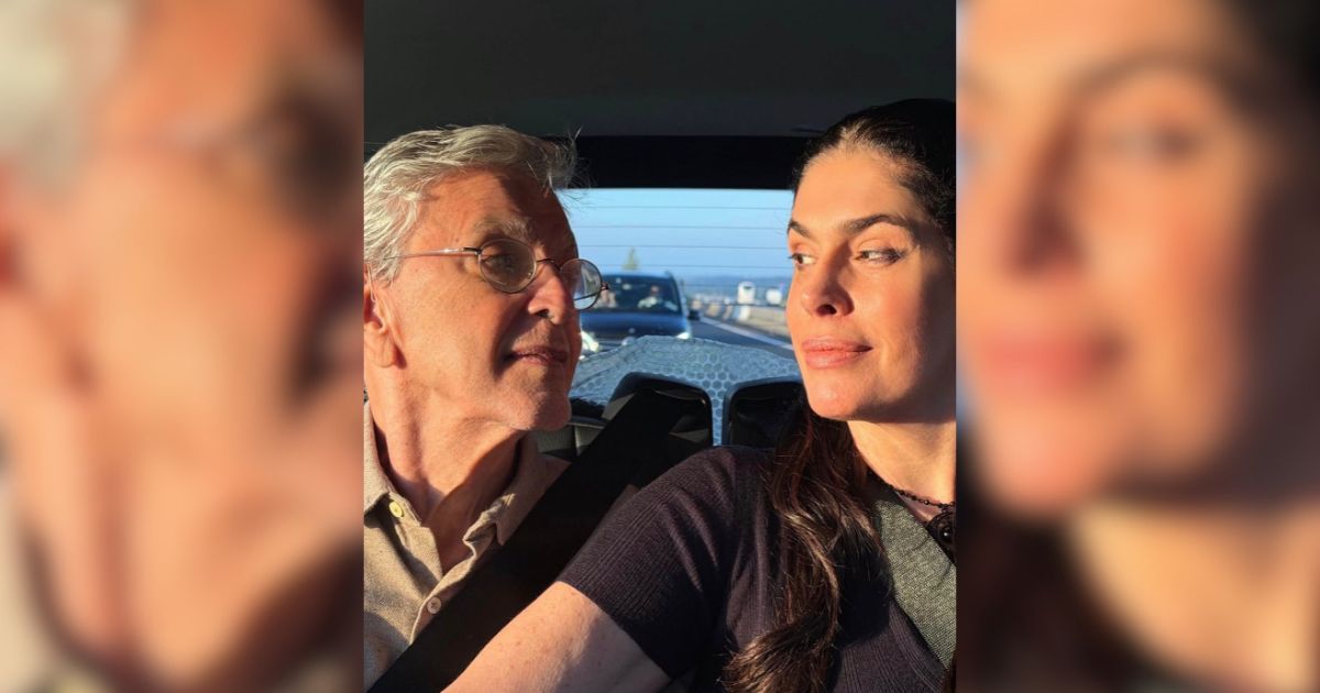 Um homem de &oacute;culos e cabelos grisalhos olha para uma mulher de cabelos escuros, ambos no banco de tr&aacute;s de um carro, com o sol iluminando seus rostos. Ao fundo, uma estrada com outros ve&iacute;culos e o c&eacute;u azul