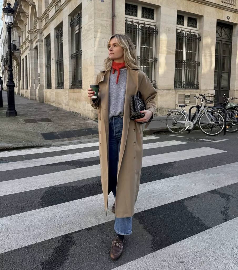 Mulher loira, com express&atilde;o s&eacute;ria, usando um sobretudo bege, su&eacute;ter cinza, jeans e len&ccedil;o vermelho, atravessa uma faixa de pedestres segurando um copo de caf&eacute; e uma bolsa preta. Ao fundo, um pr&eacute;dio antigo de pedra e bicicletas estacionadas
