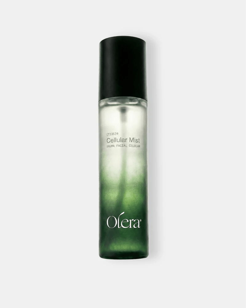 Frasco de spray de vidro transparente com tampa preta, contendo um l&iacute;quido que transita do verde escuro na base para o transl&uacute;cido no topo. O r&oacute;tulo branco exibe Cellular Mist BRUMA FACIAL CELULAR e o logo Olera em branco