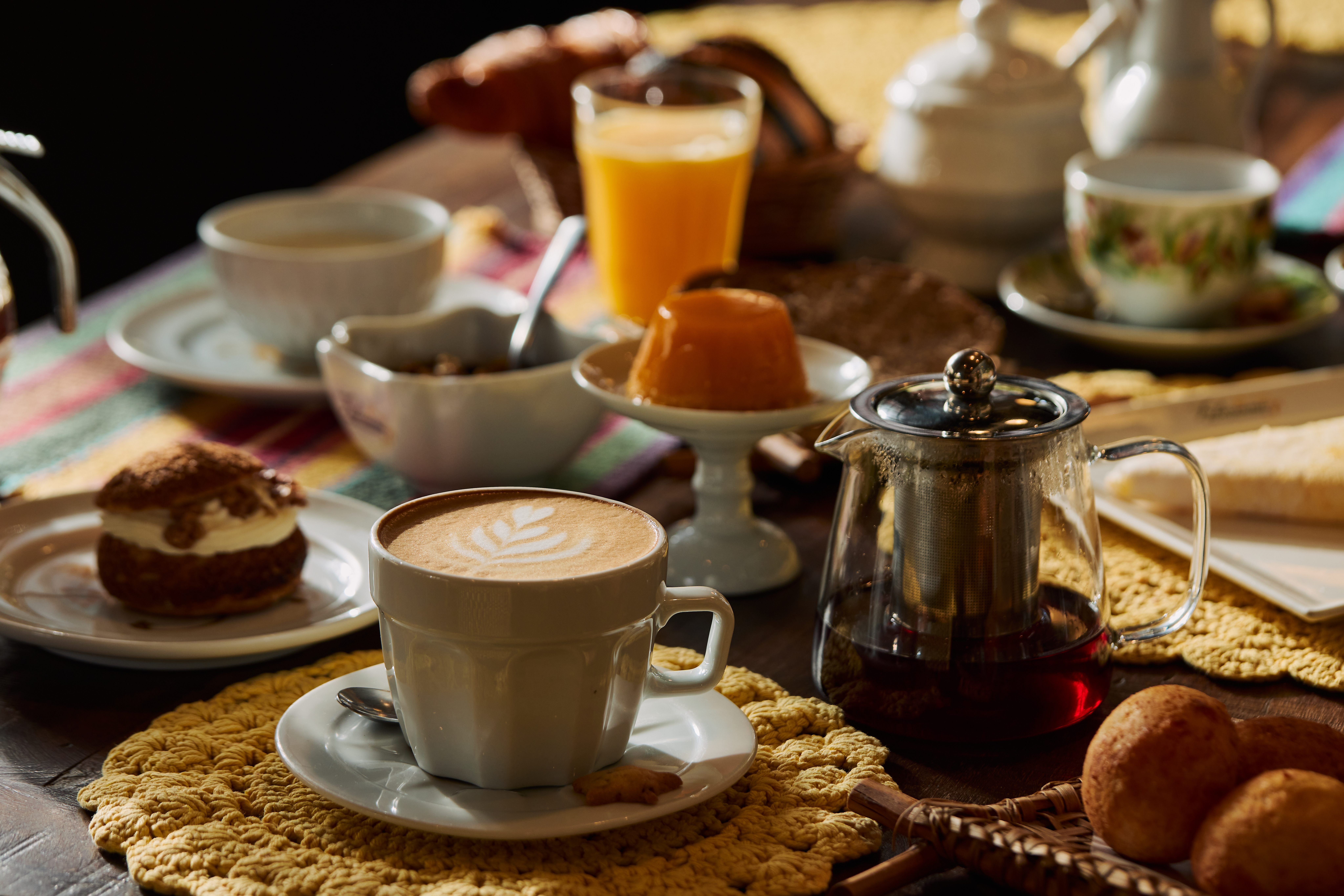 Mesa de madeira escura com caf&eacute; latte, bolo, suco de laranja, ch&aacute; e p&atilde;es, em um caf&eacute; da manh&atilde; farto