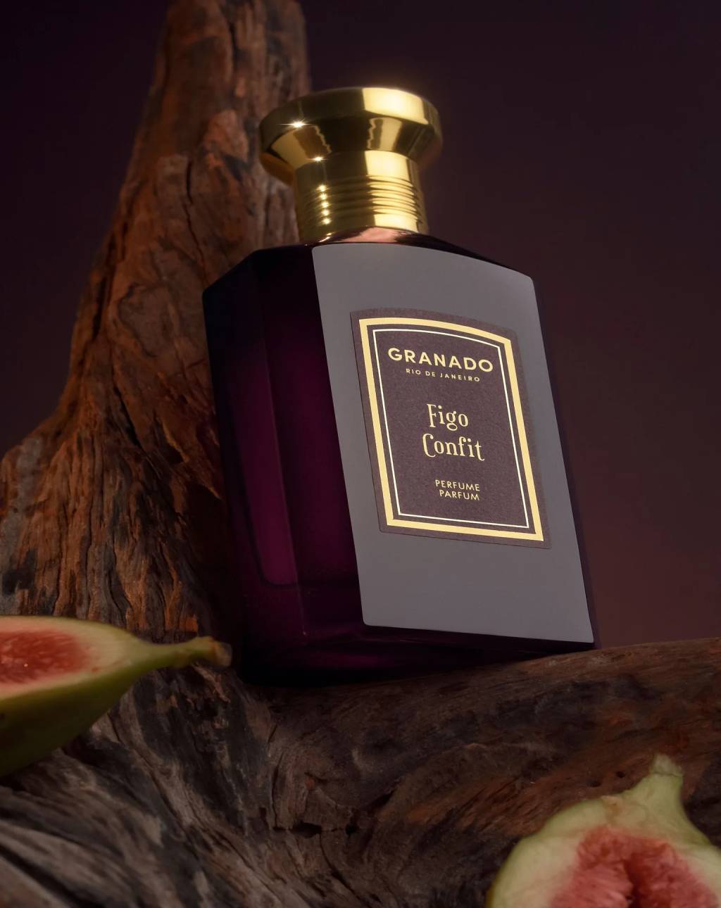 Frasco de perfume Granado Figo Confit com tampa dourada, sobre um tronco de madeira r&uacute;stica. Dois figos cortados ao meio, um em cada canto inferior, revelam a polpa rosada. Fundo escuro e suavemente iluminado
