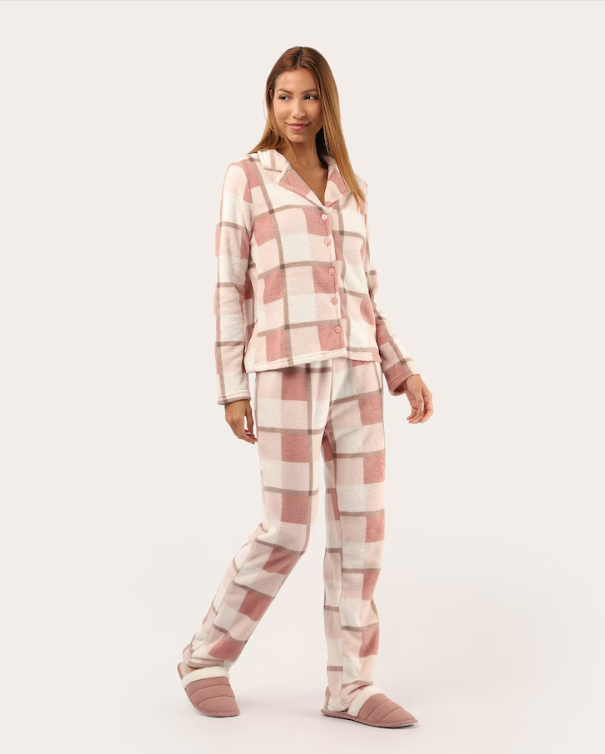 Mulher jovem, de cabelos castanhos claros e lisos, sorri levemente enquanto veste um pijama de flanela xadrez em tons de rosa claro e branco, com chinelos acolchoados da mesma cor, sobre um fundo neutro