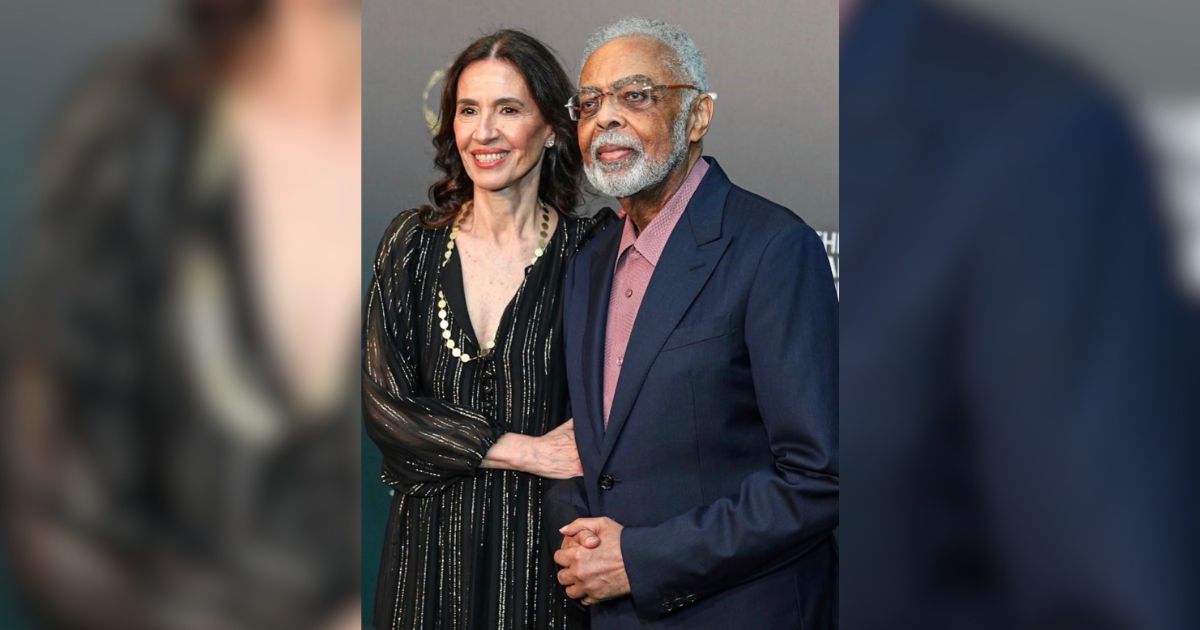 Uma mulher de cabelos escuros e um homem idoso de barba branca sorriem para a c&acirc;mera. Ela veste um vestido preto com listras finas e um colar de p&eacute;rolas, ele usa um terno azul-marinho e camisa rosa