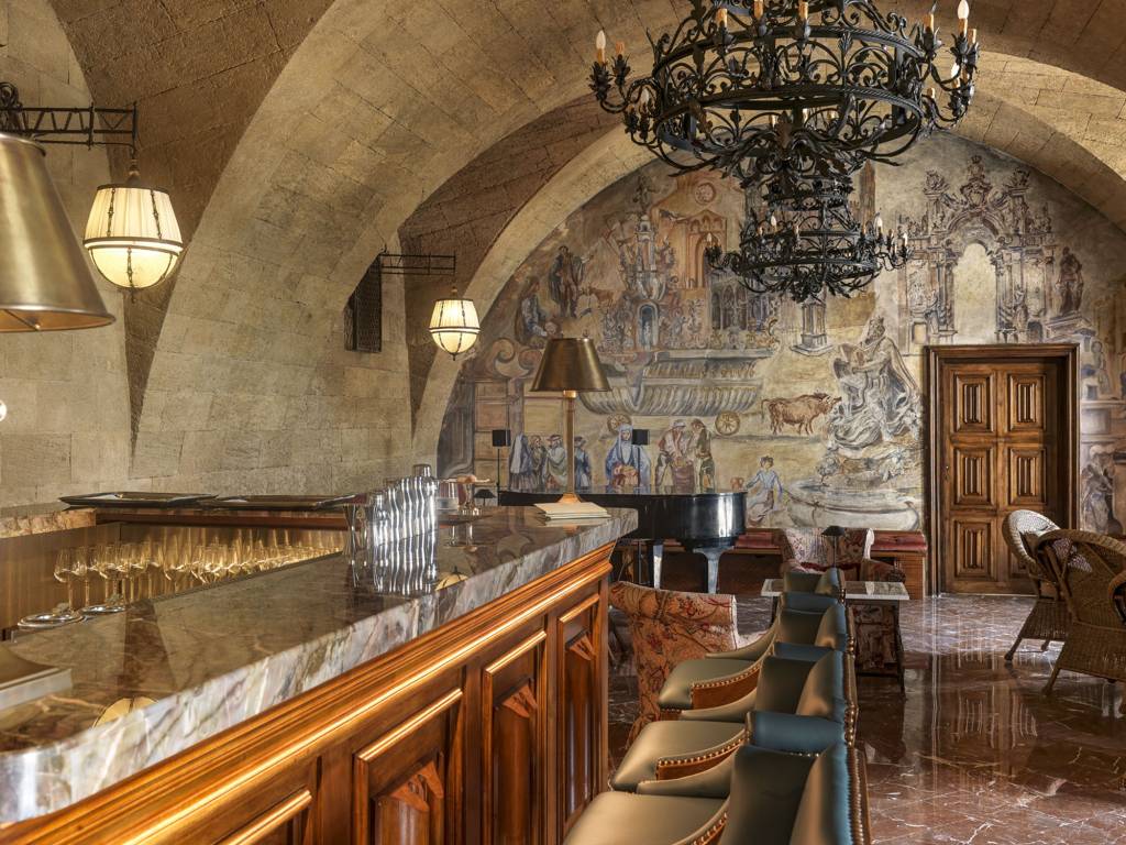 Bar luxuoso com teto abobadado de pedra, balc&atilde;o de m&aacute;rmore e madeira, ta&ccedil;as de vinho alinhadas. Ao fundo, uma parede com afrescos detalhados de cenas hist&oacute;ricas, um piano de cauda, cadeiras estofadas e lustres de ferro forjado pendurados. O piso &eacute; de m&aacute;rmore marrom brilhante e uma porta de madeira entalhada se destaca na parede pintada