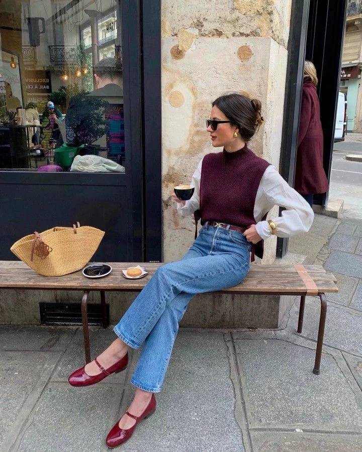 Mulher sentada em banco de madeira, vestindo camisa branca, colete vinho, jeans e sapatilhas vermelhas. Ela segura uma x&iacute;cara de caf&eacute;, com &oacute;culos escuros e brincos dourados. Ao lado, uma bolsa de palha e um doce.