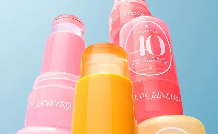Tr&ecirc;s frascos de perfume em bast&atilde;o, dois rosa e um laranja, com o nome "Sol de Janeiro" e "Cheirosa 40 Jelly Perfume Balm" em um fundo azul claro