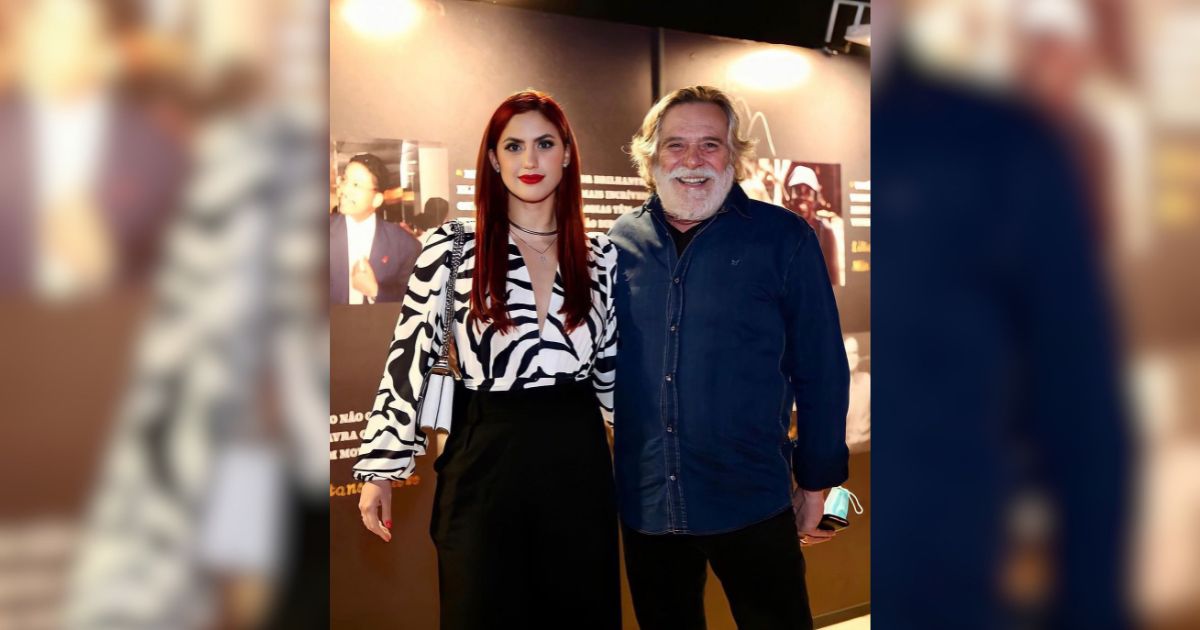 Uma mulher de cabelos ruivos e blusa zebra preta e branca, ao lado de um homem grisalho com barba e camisa jeans azul, ambos sorrindo para a c&acirc;mera em um evento noturno