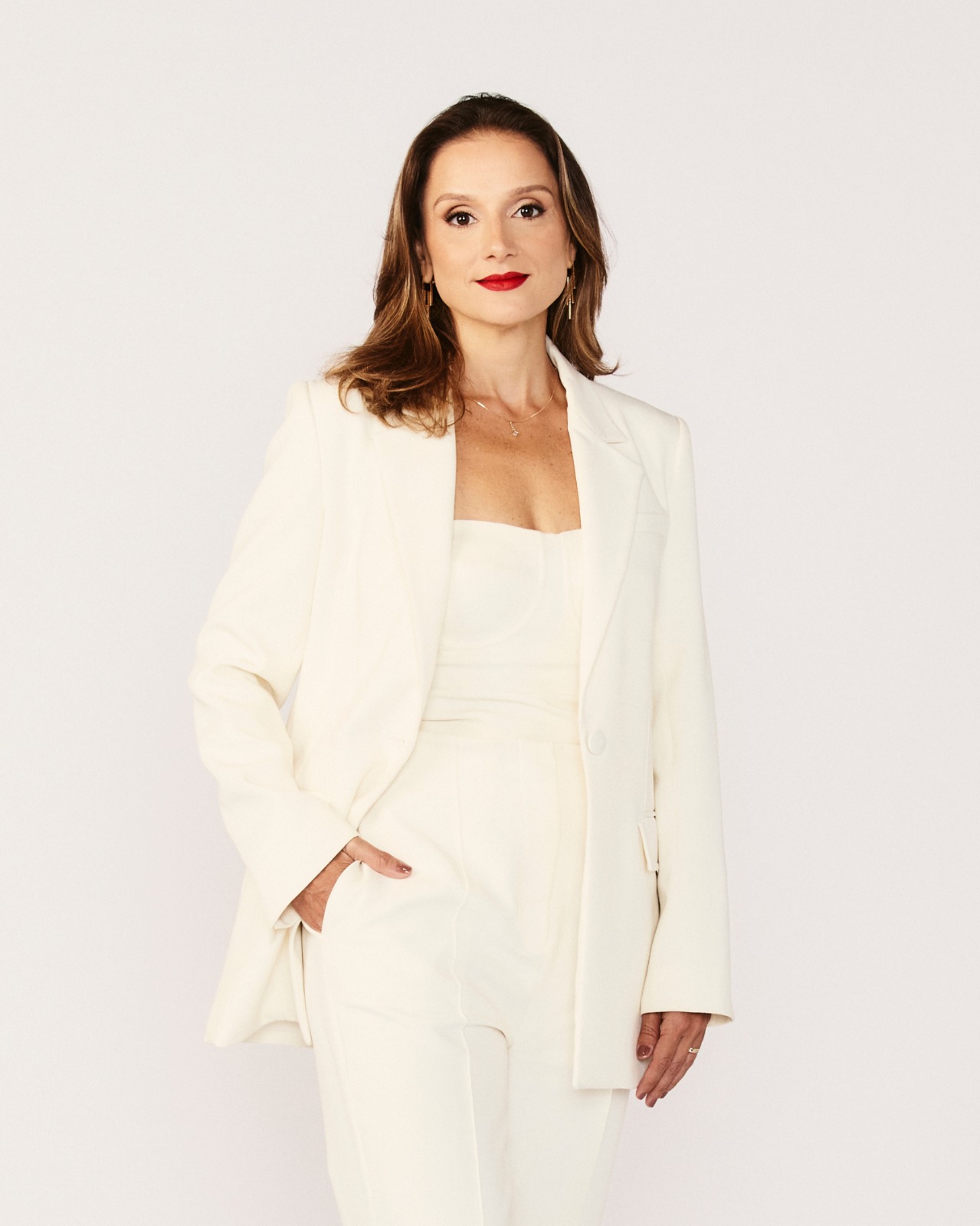 Mulher de cabelos castanhos claros, maquiagem com batom vermelho, vestindo blazer e cal&ccedil;a creme, com uma m&atilde;o no bolso e a outra relaxada ao lado do corpo, em fundo branco