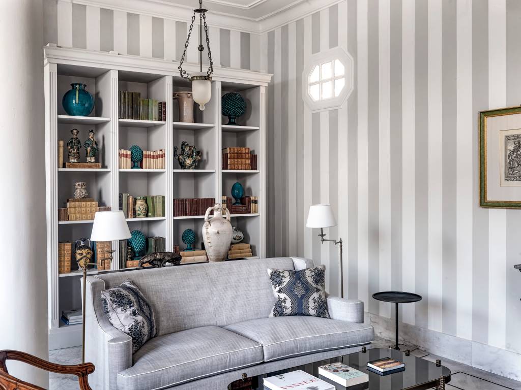 Sala de estar com sof&aacute; cinza, almofadas estampadas, estante branca cheia de livros e objetos decorativos, e paredes com listras verticais cinza e brancas