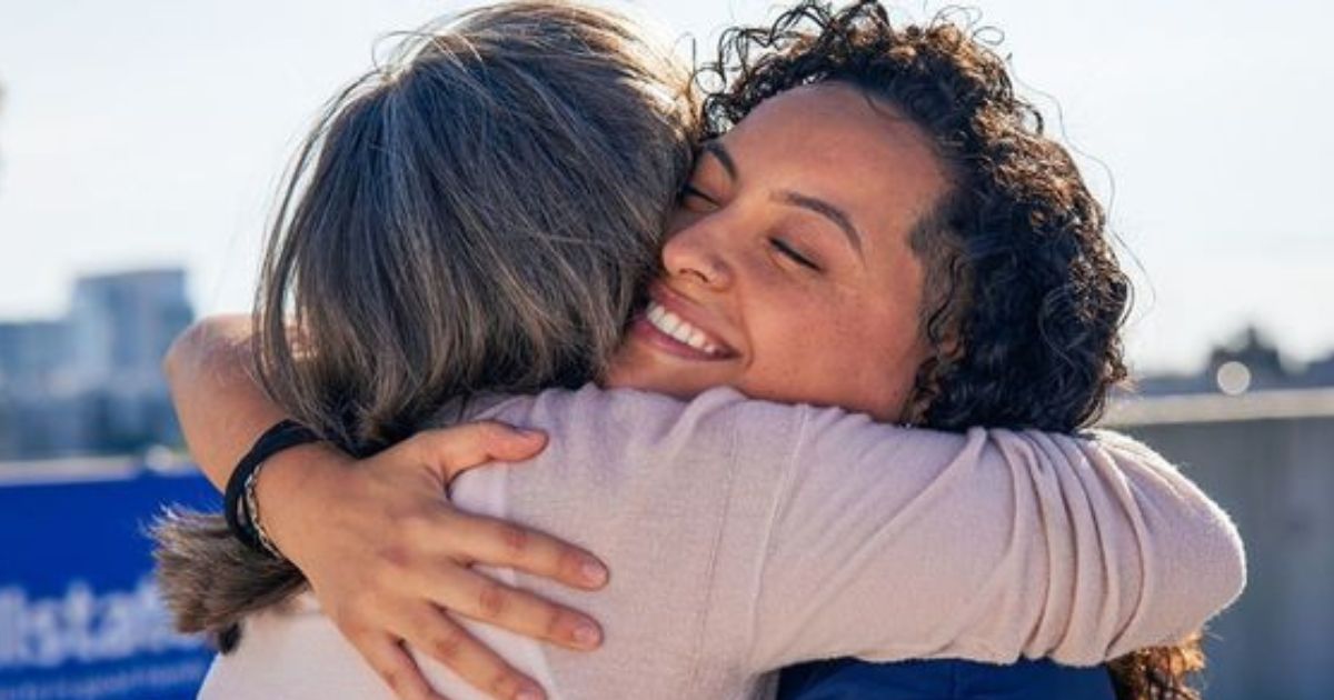 Mulher de cabelos cacheados e escuros, sorrindo com os olhos fechados, abra&ccedil;a outra pessoa de costas, com cabelos grisalhos. A cena transmite afeto e felicidade, com um fundo desfocado de c&eacute;u claro e edif&iacute;cios