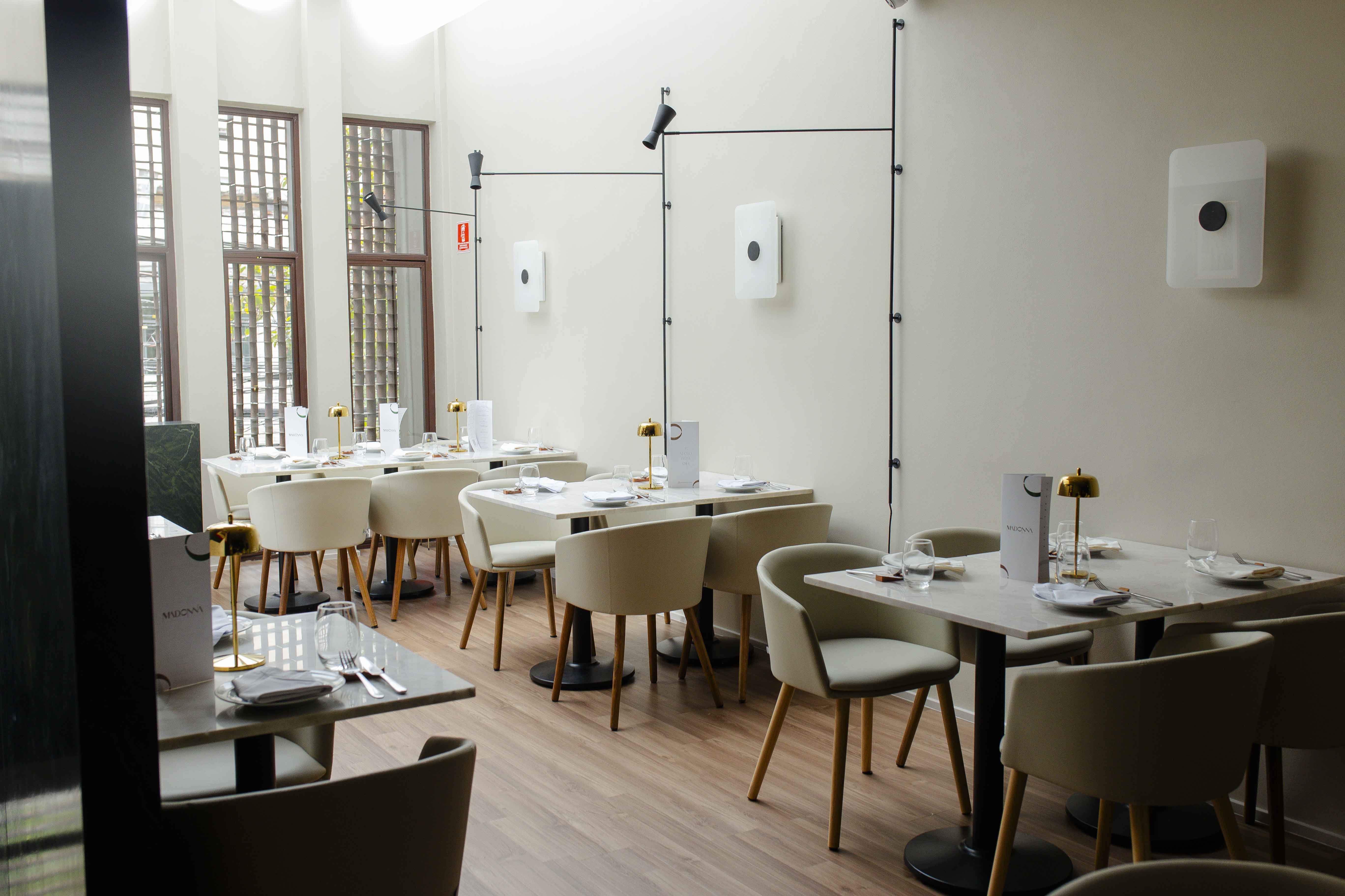 Interior de restaurante moderno com mesas de m&aacute;rmore branco, cadeiras estofadas bege e piso de madeira. As mesas est&atilde;o postas com pratos, talheres, copos e lumin&aacute;rias douradas. A parede clara tem janelas altas com treli&ccedil;as de madeira e lumin&aacute;rias pretas com focos direcion&aacute;veis