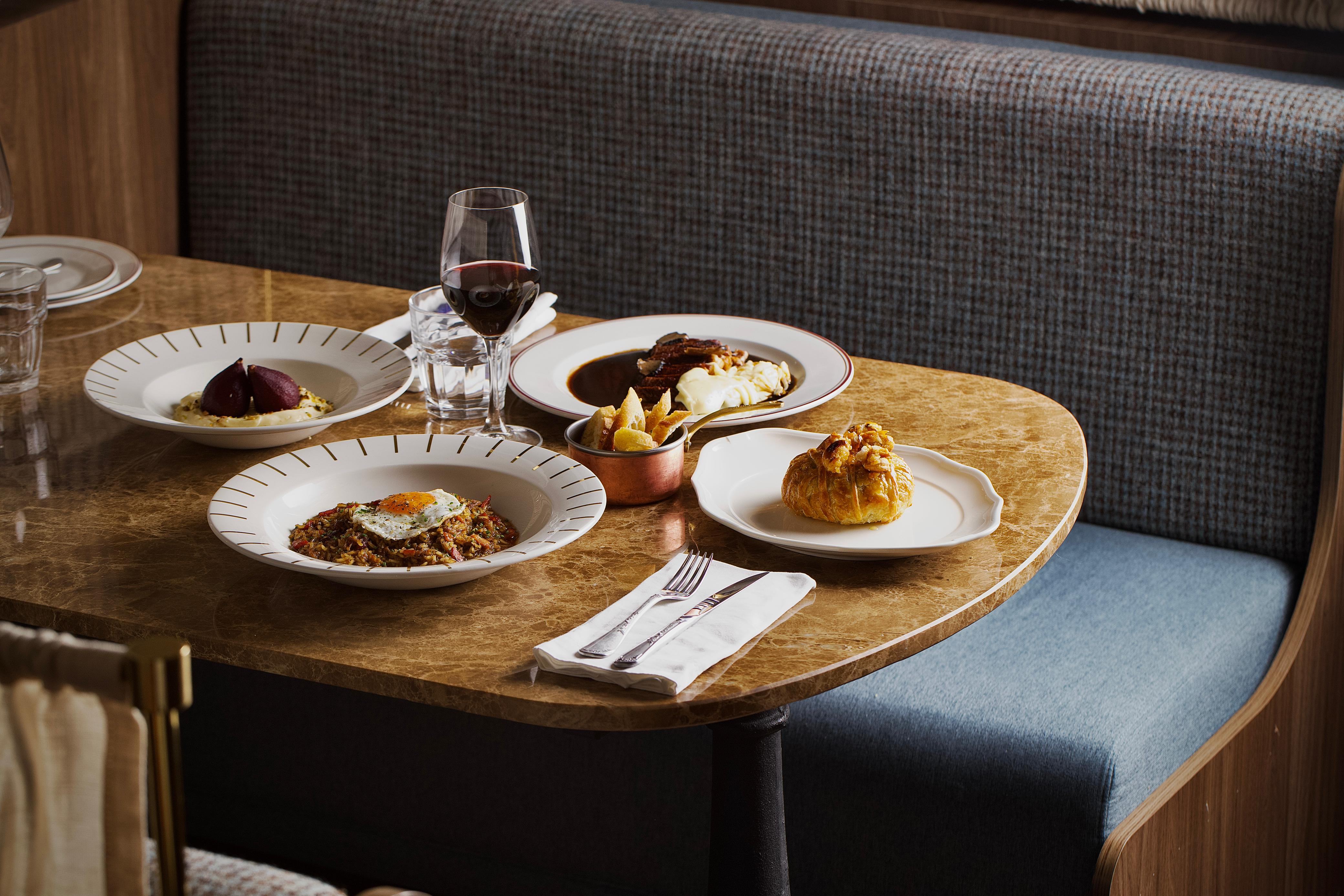 Mesa de restaurante com pratos gourmet: um com arroz e ovo frito, outro com peras e pur&ecirc;, um terceiro com carne e pur&ecirc;, um p&atilde;ozinho folhado e batatas fritas, al&eacute;m de um copo de vinho tinto e talheres sobre guardanapo branco