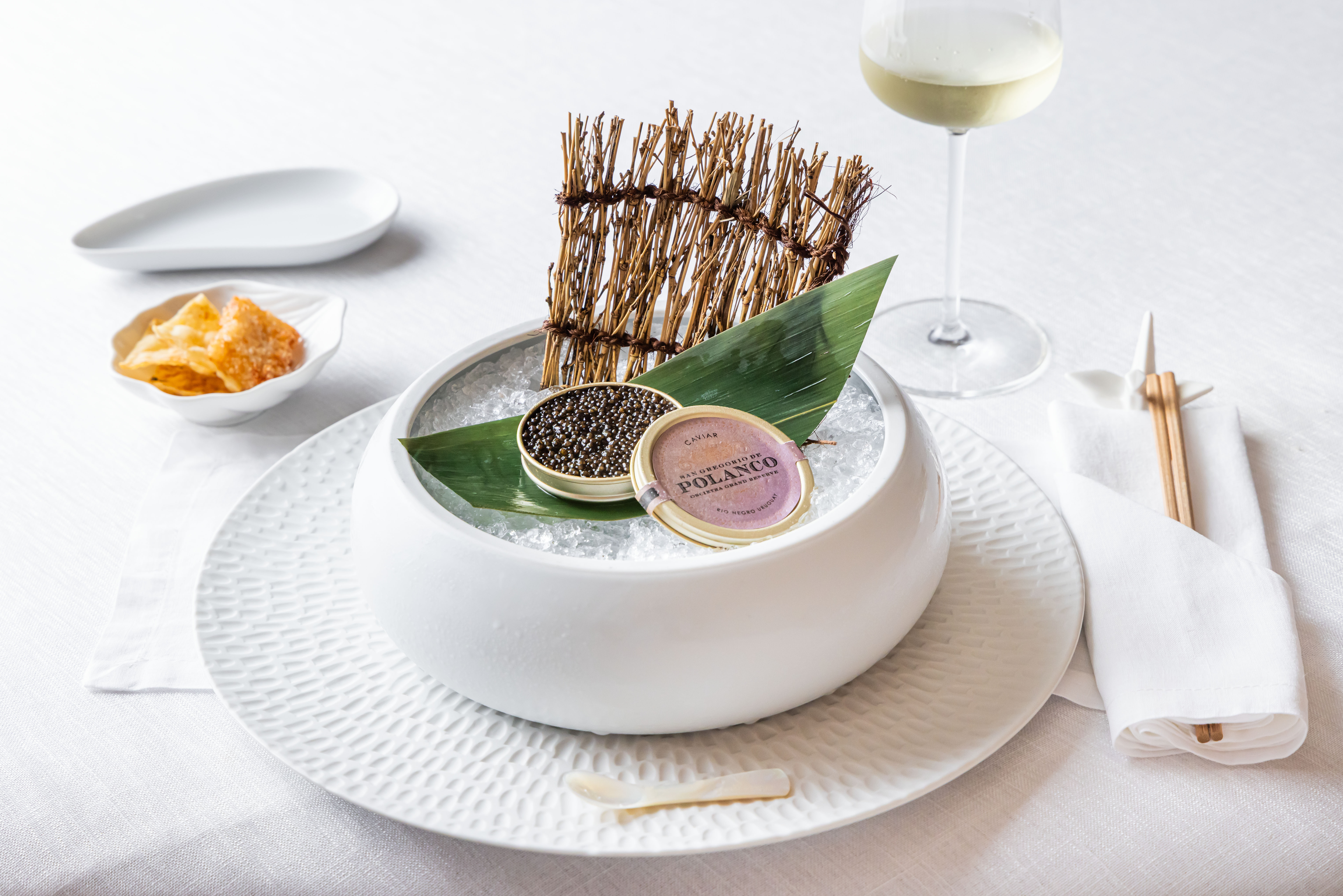Uma tigela branca com gelo e caviar Polanco, acompanhada de batatas chips, um copo de vinho branco e hashi, sobre uma toalha de mesa branca
