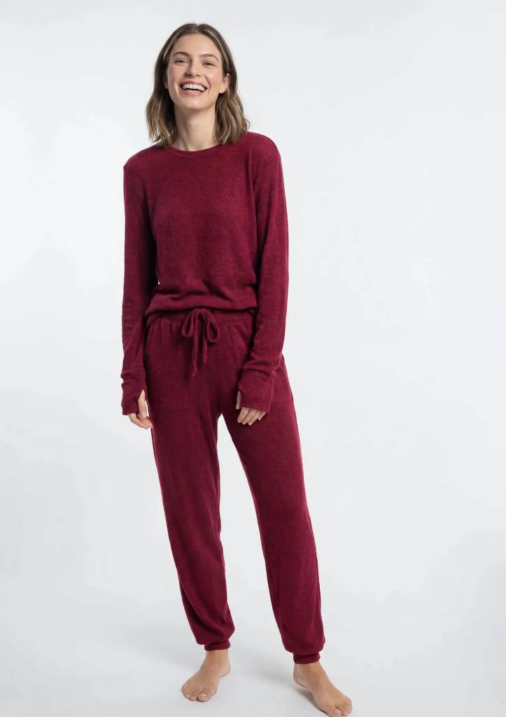 Mulher de pele clara, cabelos castanhos curtos e sorriso largo, vestindo um conjunto de moletom felpudo cor vinho, com blusa de manga comprida e cal&ccedil;a jogger com amarra&ccedil;&atilde;o na cintura e bolsos, descal&ccedil;a, em fundo branco
