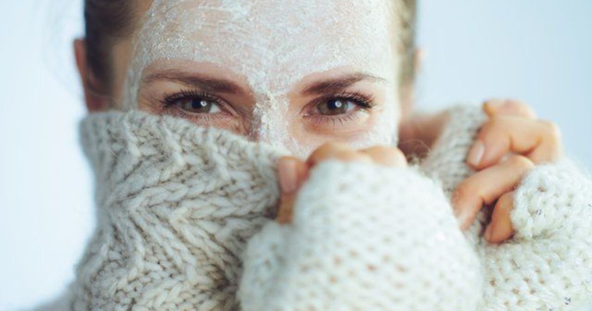 Mulher com m&aacute;scara facial branca no rosto, puxando a gola de um su&eacute;ter de tric&ocirc; creme para cobrir o nariz e a boca, olhando para frente