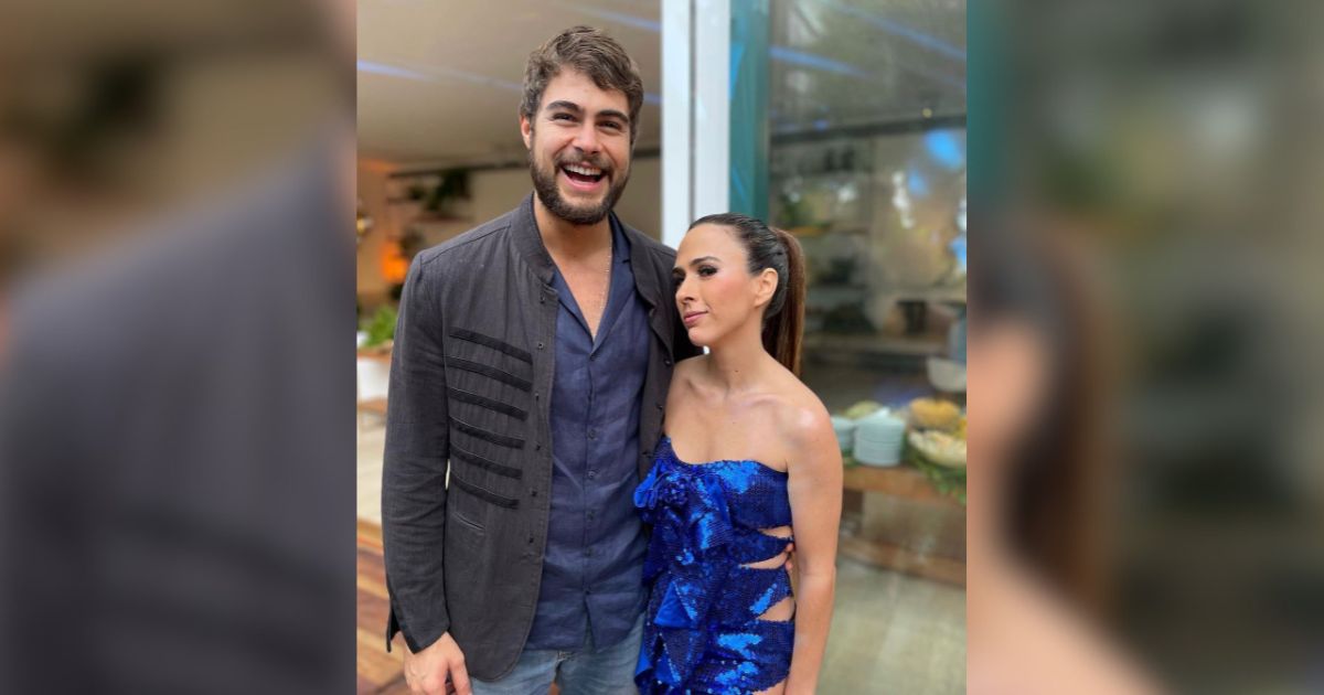 Um homem barbudo sorrindo amplamente, vestindo camisa azul e jaqueta preta, ao lado de uma mulher com rabo de cavalo e vestido azul brilhante, olhando para ele com carinho