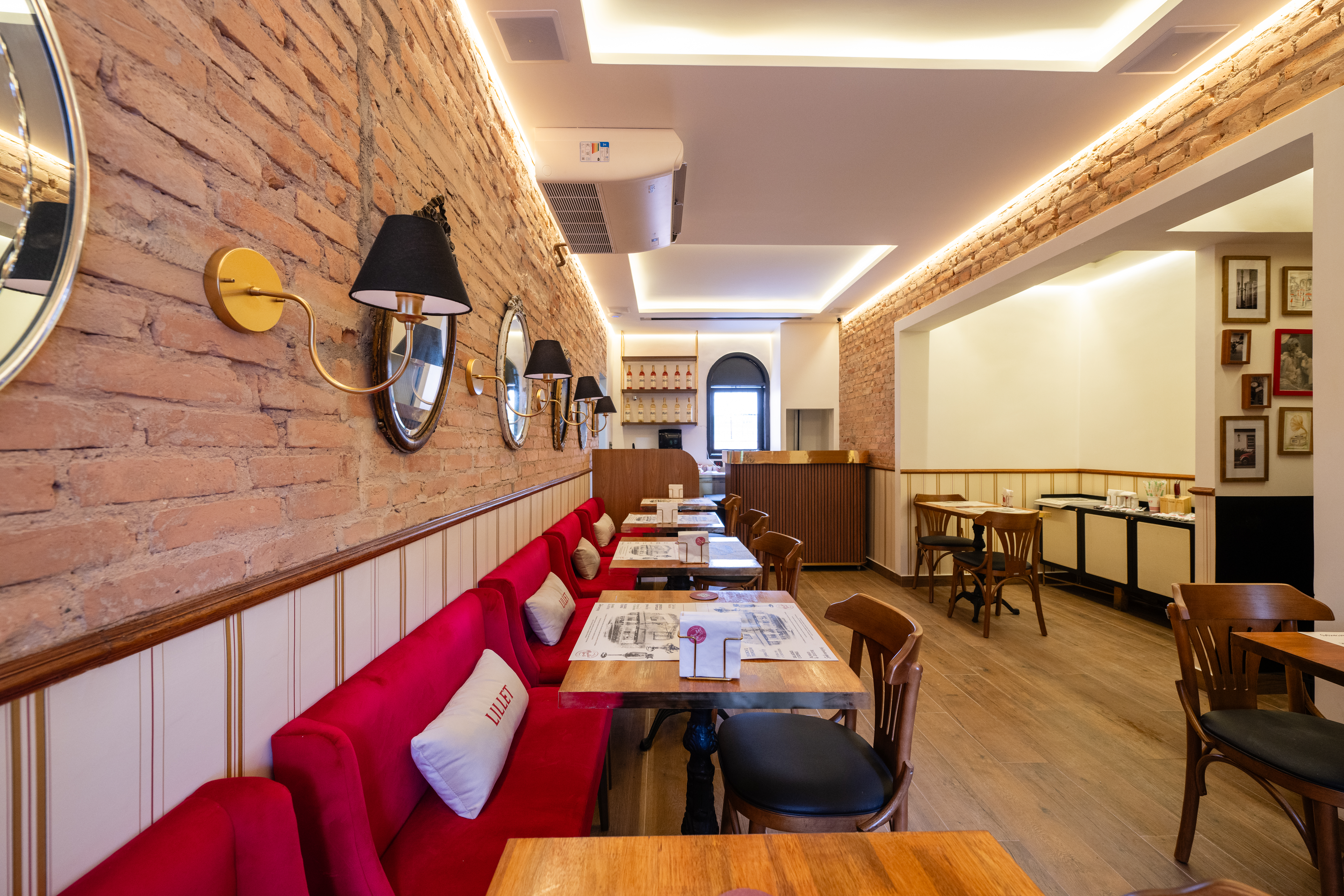 Interior de um restaurante com paredes de tijolos expostos e bancos estofados vermelhos. Mesas de madeira com cadeiras escuras, espelhos ovais e arandelas douradas com c&uacute;pulas pretas decoram o ambiente. Ao fundo, um balc&atilde;o de madeira e uma porta arqueada.