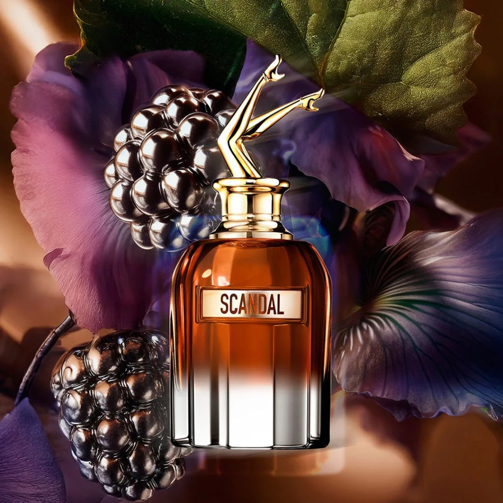 Frasco de perfume Scandal com l&iacute;quido &acirc;mbar e base transparente, tampa dourada com pernas femininas cruzadas, sobre fundo de folhas roxas e amoras escuras