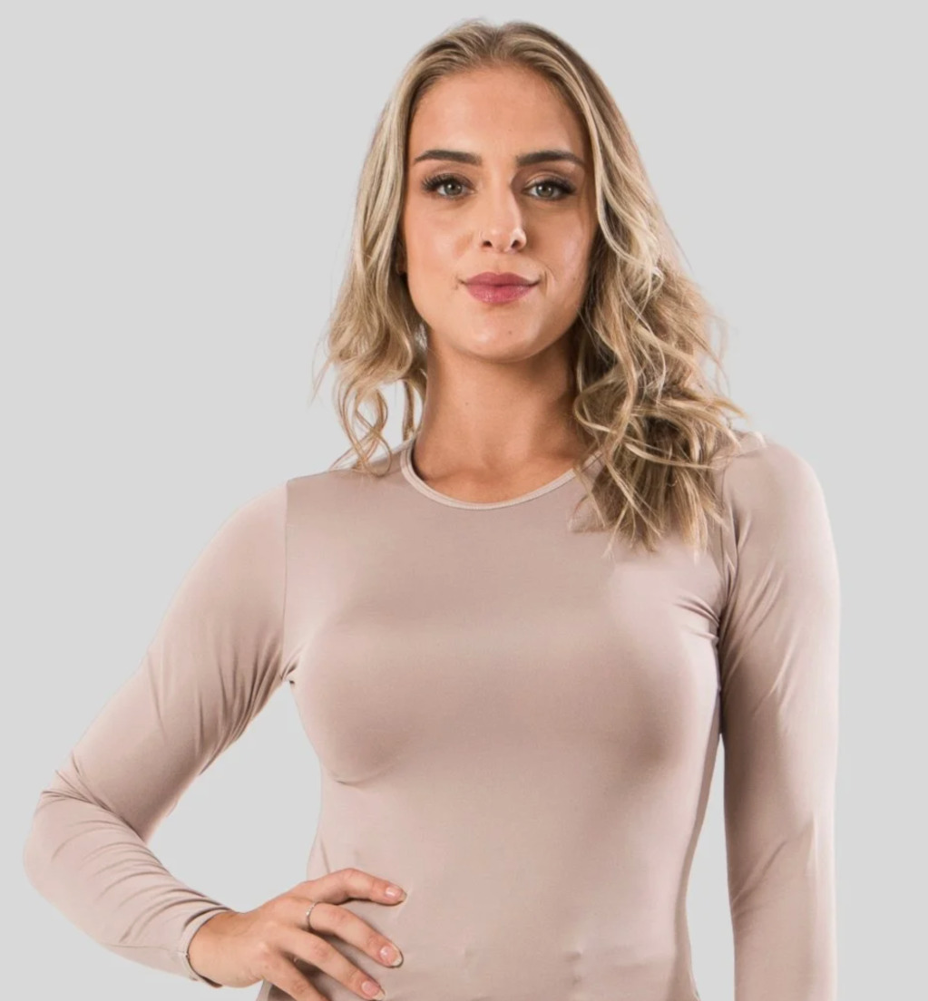 Mulher loira de cabelo ondulado, vestindo blusa bege de manga comprida, com a m&atilde;o esquerda na cintura e express&atilde;o s&eacute;ria, em fundo cinza